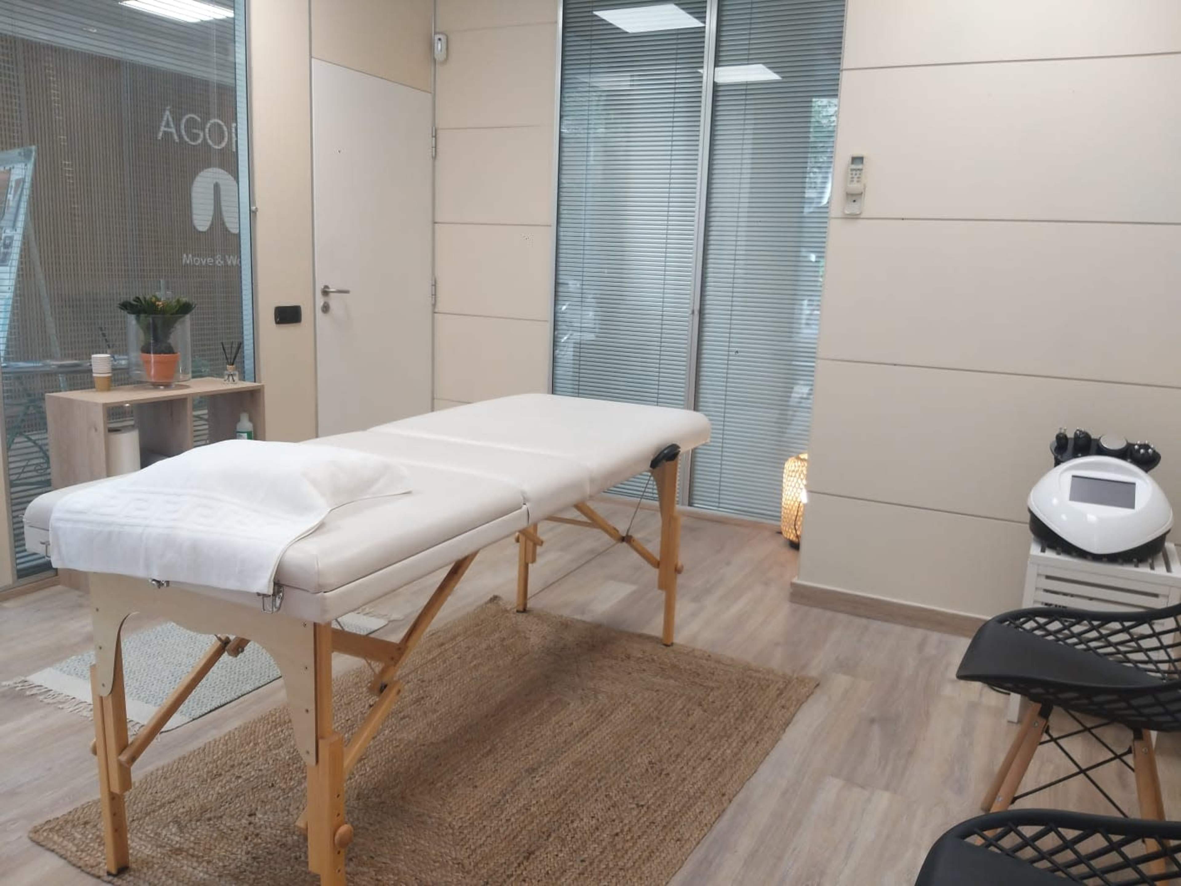 A massage therapy room with a white massage table, wood flooring, and large glass windows.