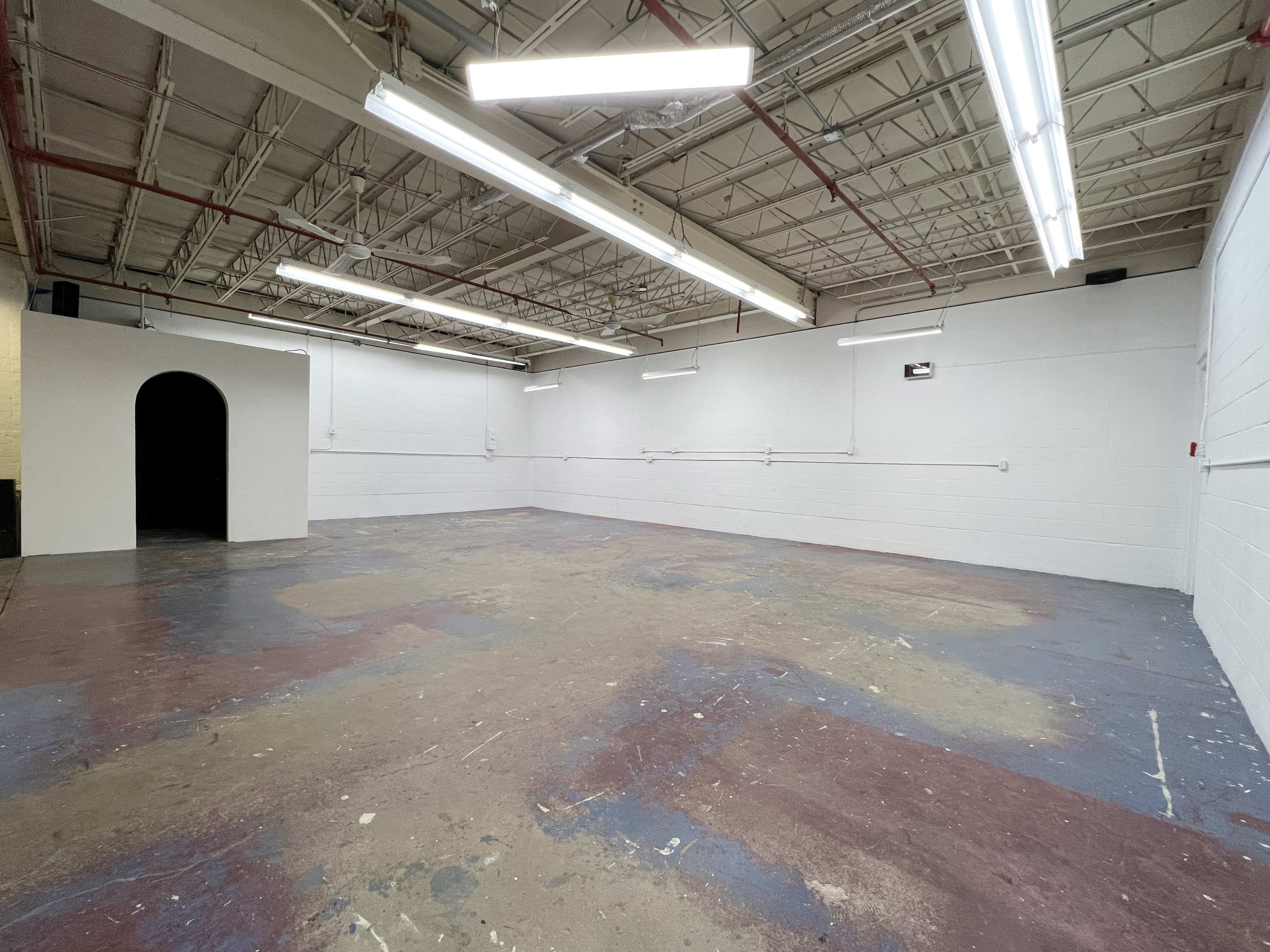 A spacious, empty interior room with concrete flooring and ceiling lights, featuring a dark archway on the left side.