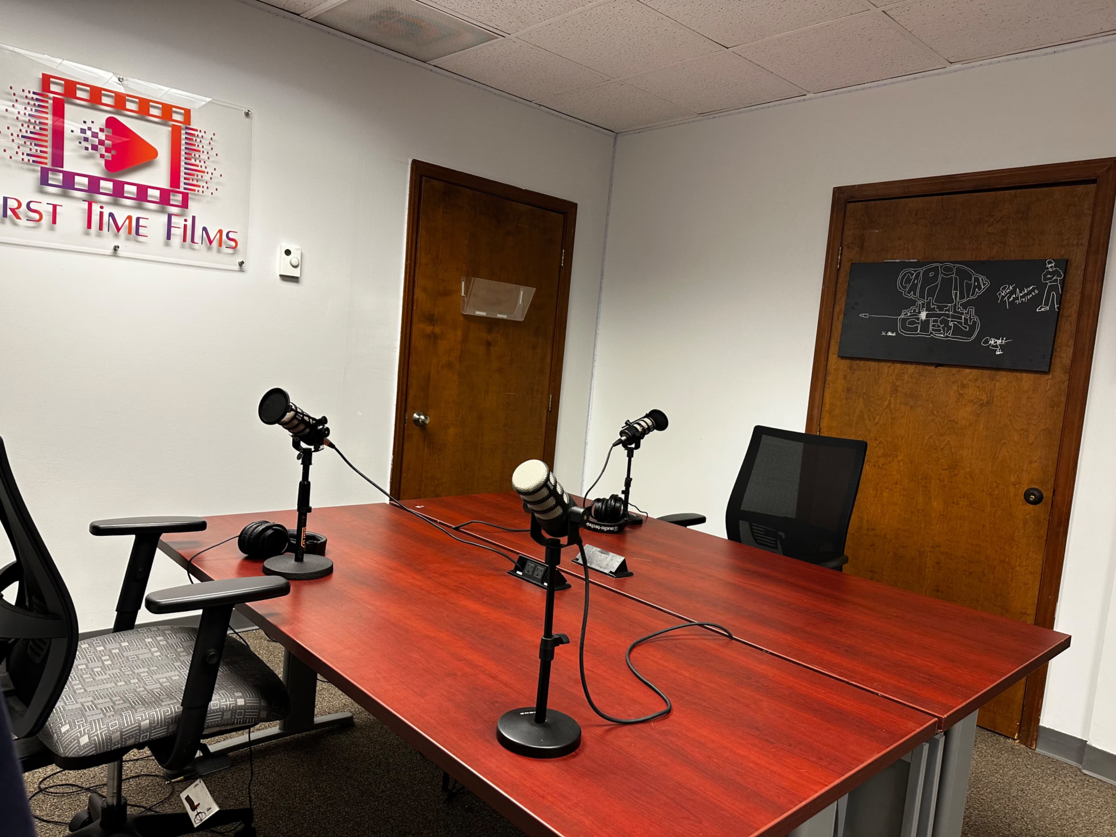 The image shows a small recording studio with a red table, three microphones, and a blackboard on the wall, alongside wooden doors and a sign that reads "First Time Films."