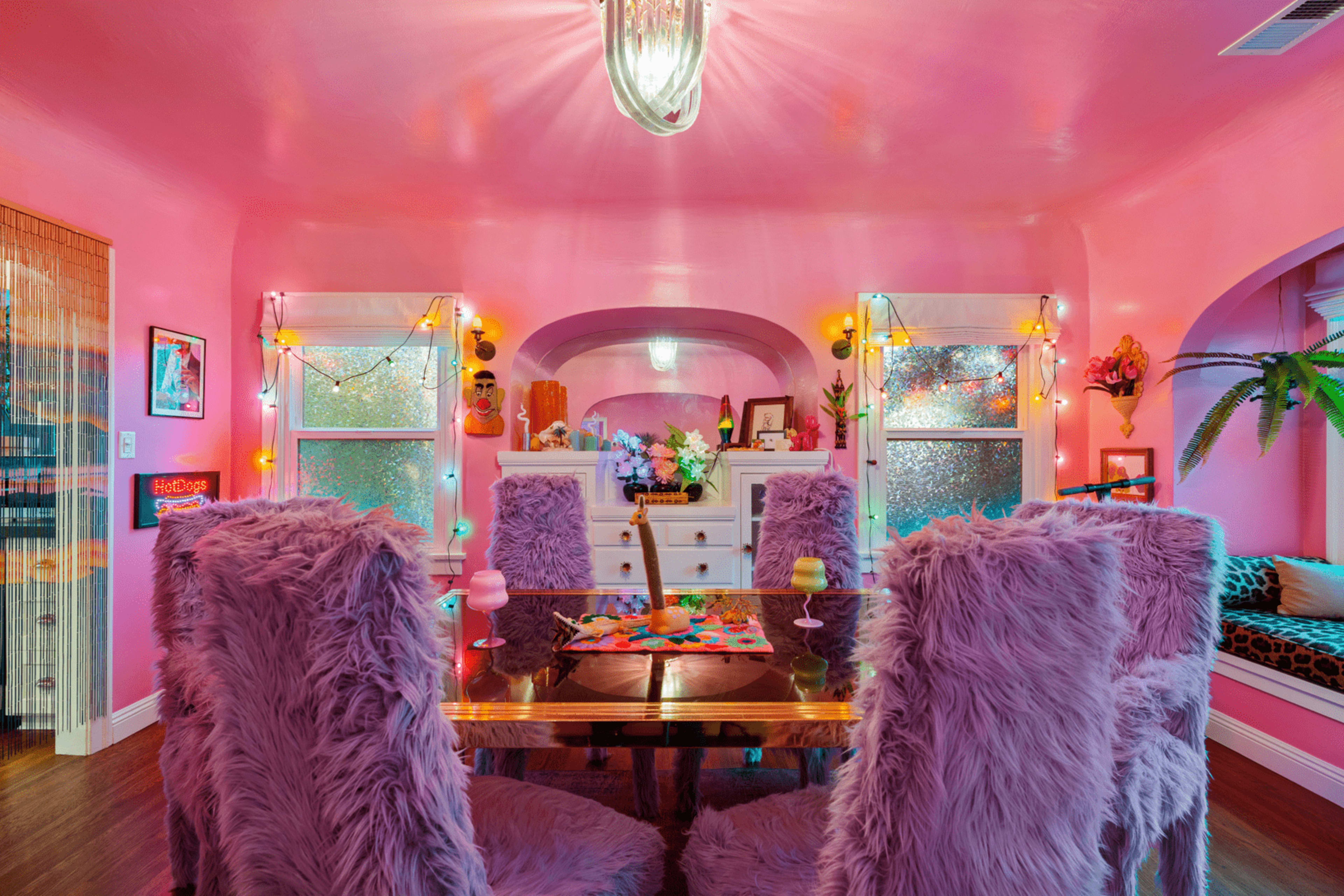 A pink-themed dining area features a table surrounded by fluffy chairs, illuminated by decorative lights and a central pendant fixture.