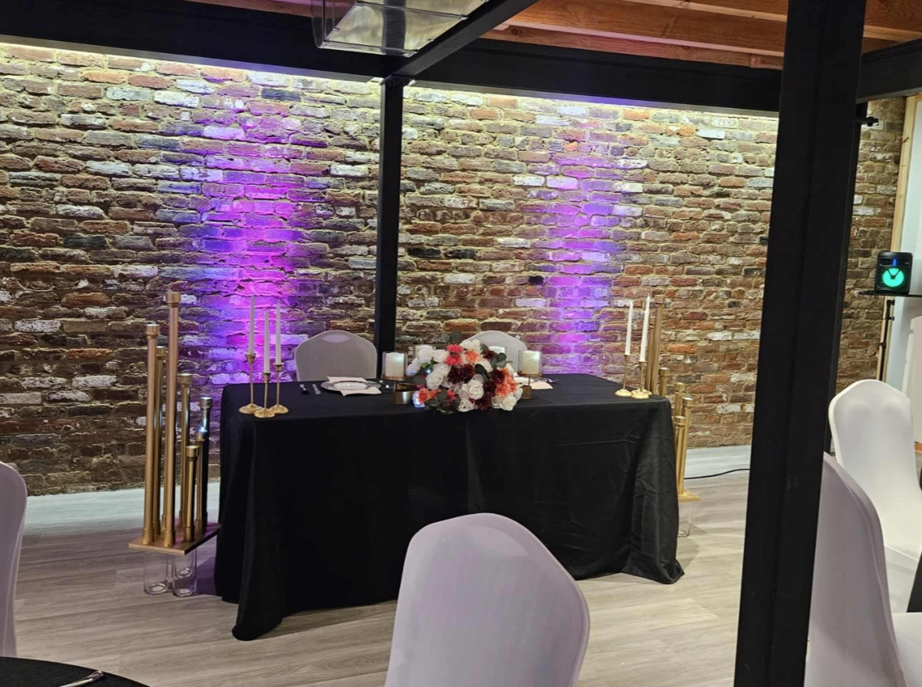 A table set with a black tablecloth, decorated with white and orange flowers, candles, and positioned against a brick wall illuminated with purple light.
