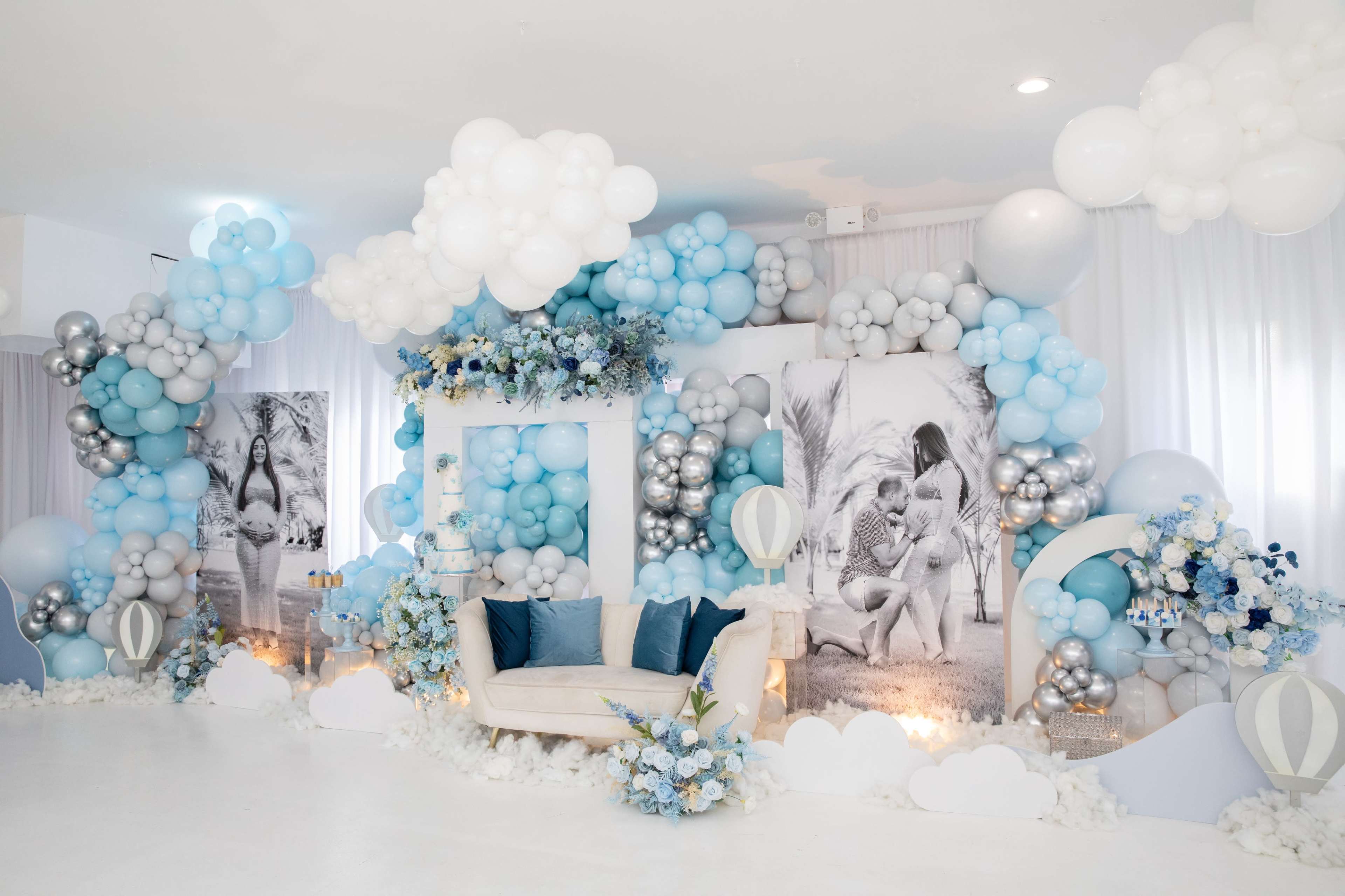 The image shows a decorated event space with a couch surrounded by a backdrop of balloons in shades of blue and silver, along with large monochrome photos and floral arrangements.