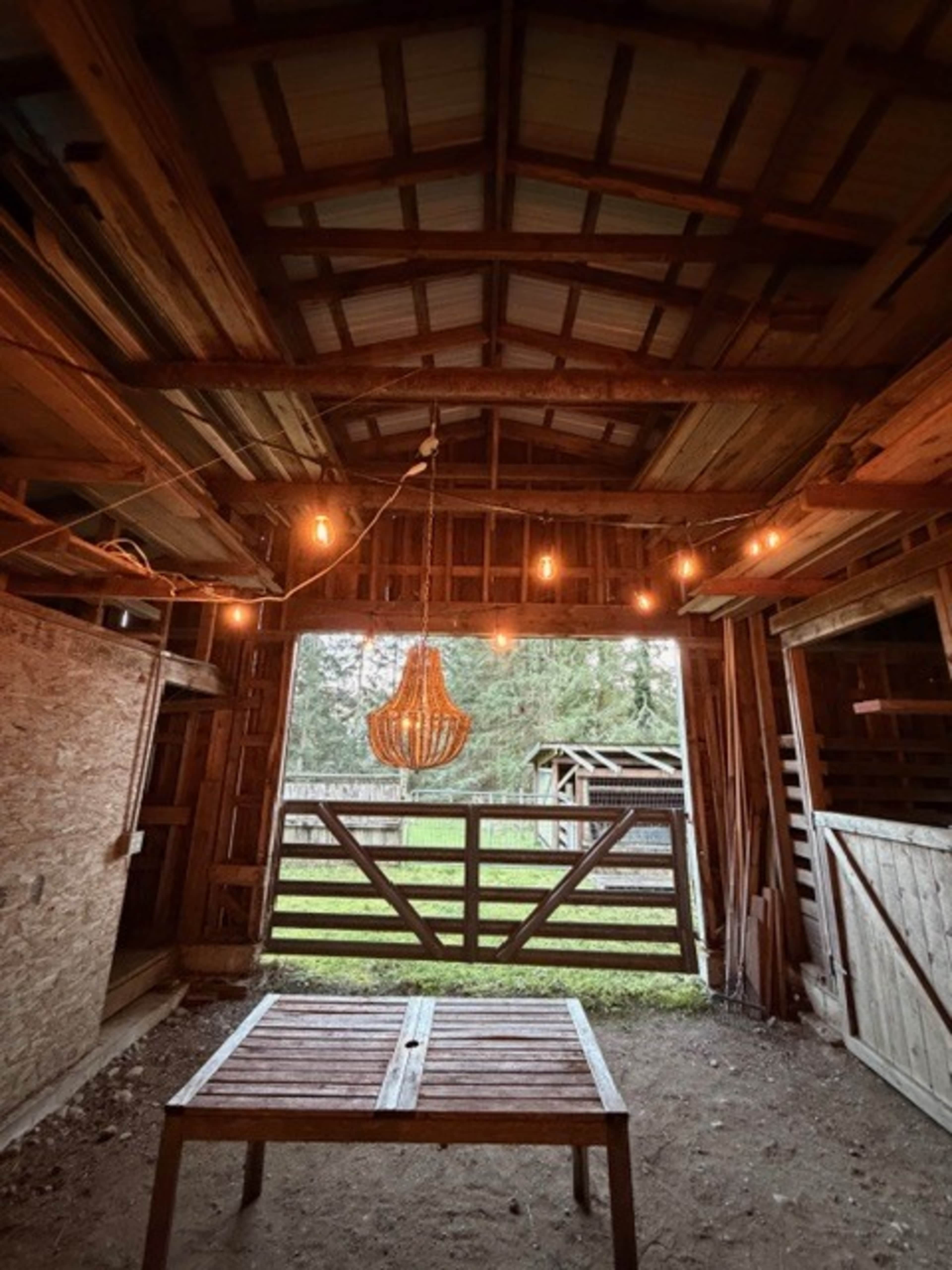 The image shows the interior of a rustic barn with wooden beams, a hanging light fixture, and a view of a fenced area outside.