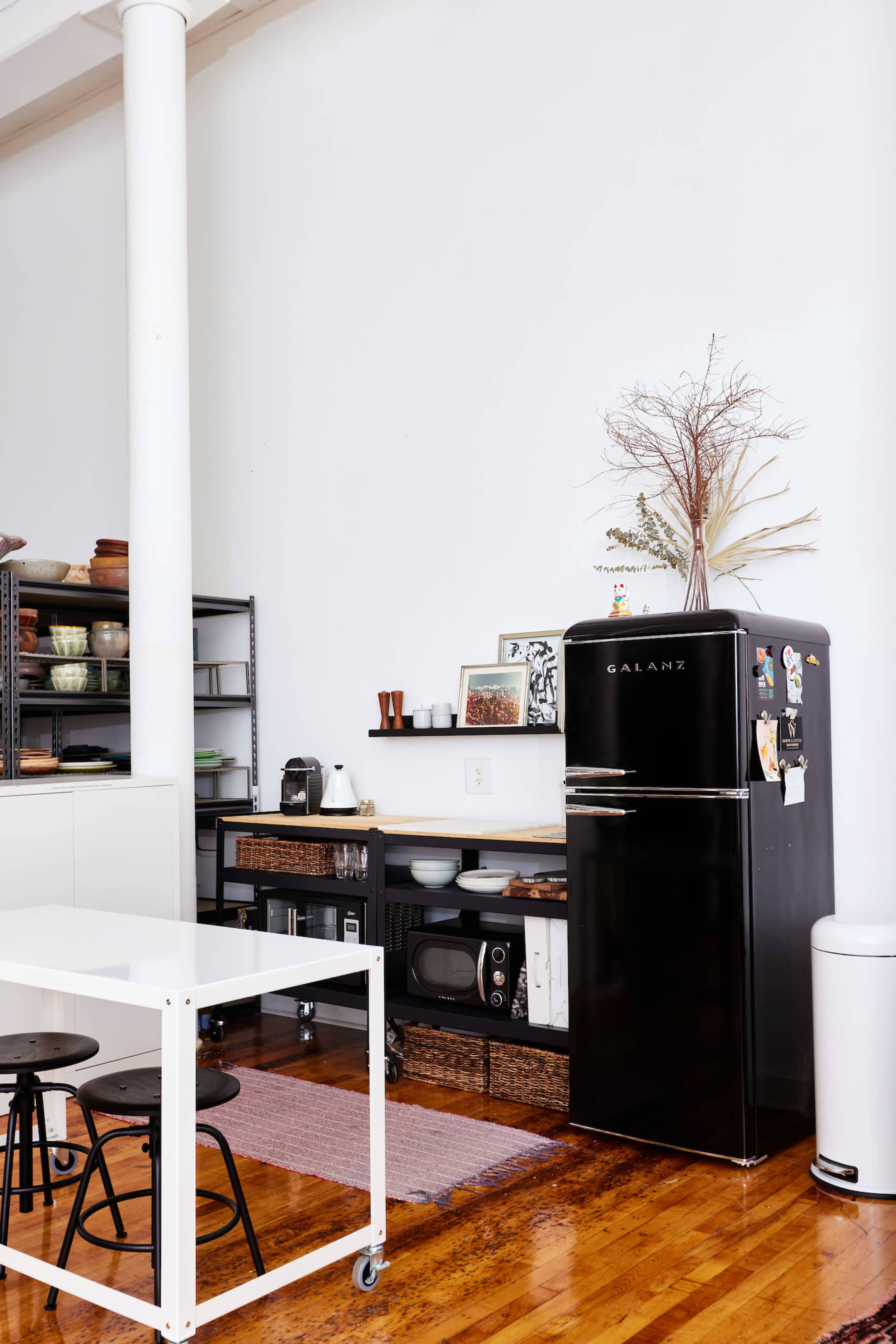 A black refrigerator is positioned next to a shelf with various kitchen items, while a white table and chairs are arranged nearby on a wooden floor.