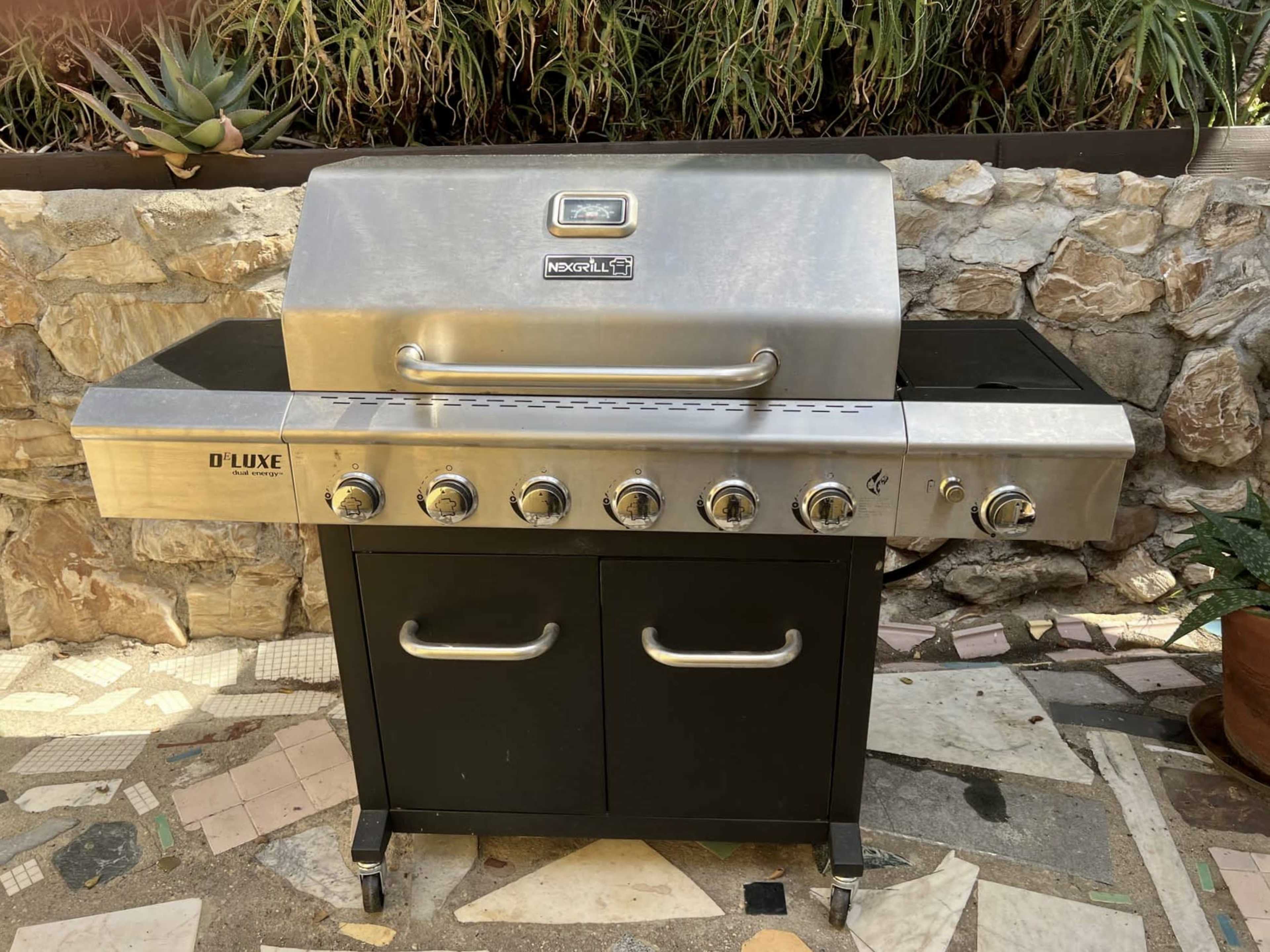 A stainless steel gas grill with a black cabinet and multiple control knobs, positioned on a stone patio surrounded by greenery.
