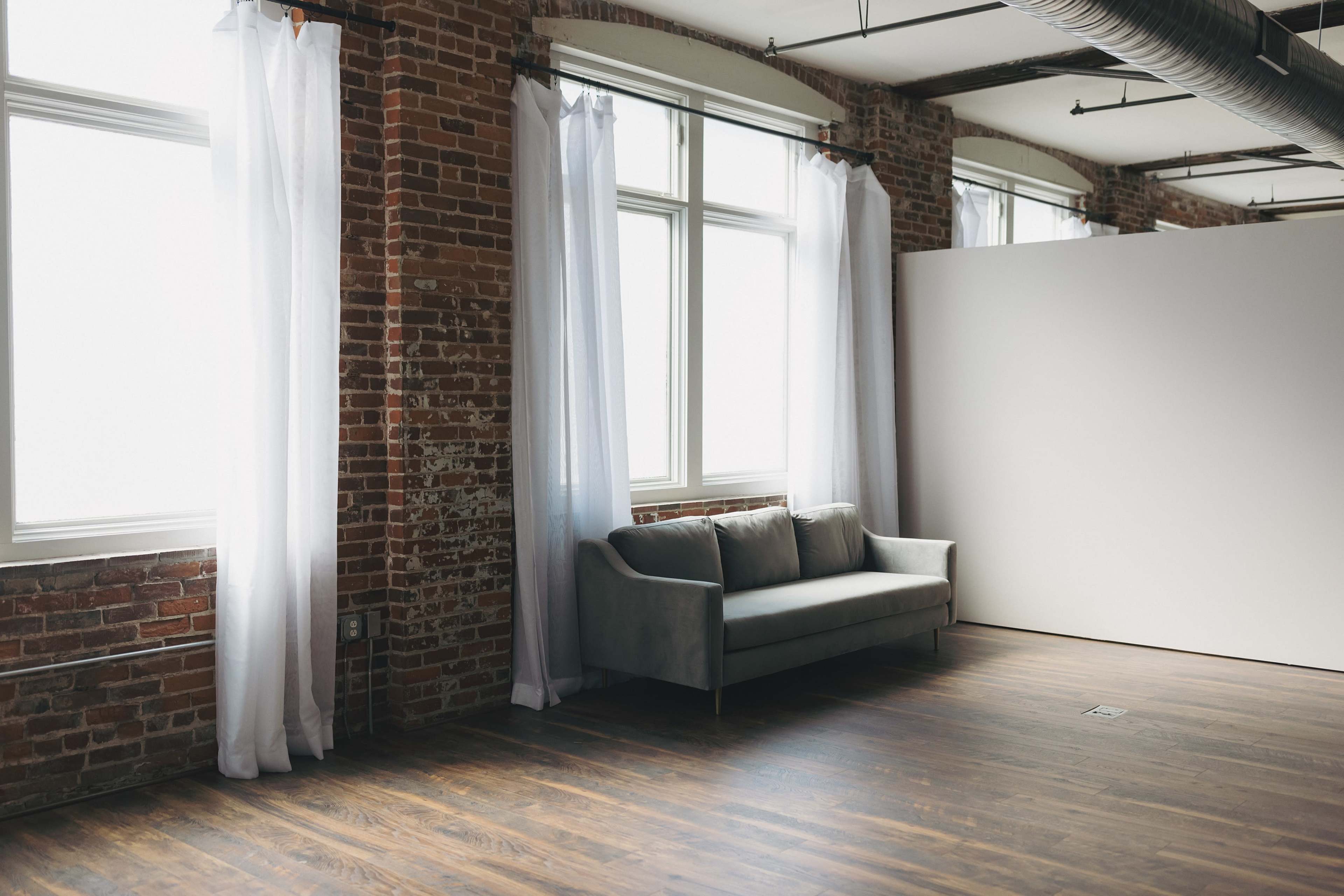 The image shows a minimalist interior space with exposed brick walls, large windows with sheer white curtains, and a gray couch positioned near a blank white partition.