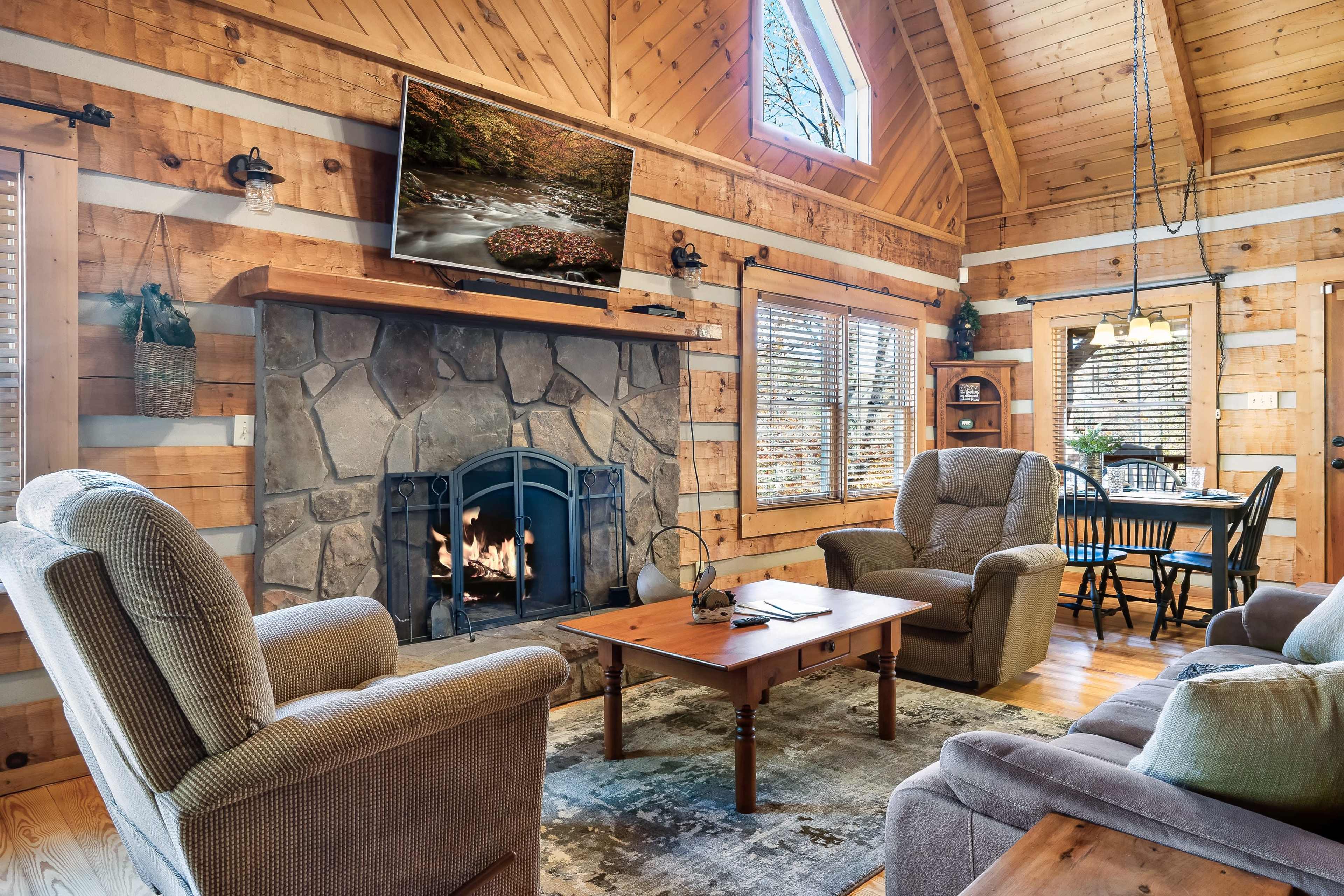 The image shows a cozy cabin living room featuring a stone fireplace, wooden walls, a television mounted above the mantel, and comfortable seating arrangements around a central coffee table.