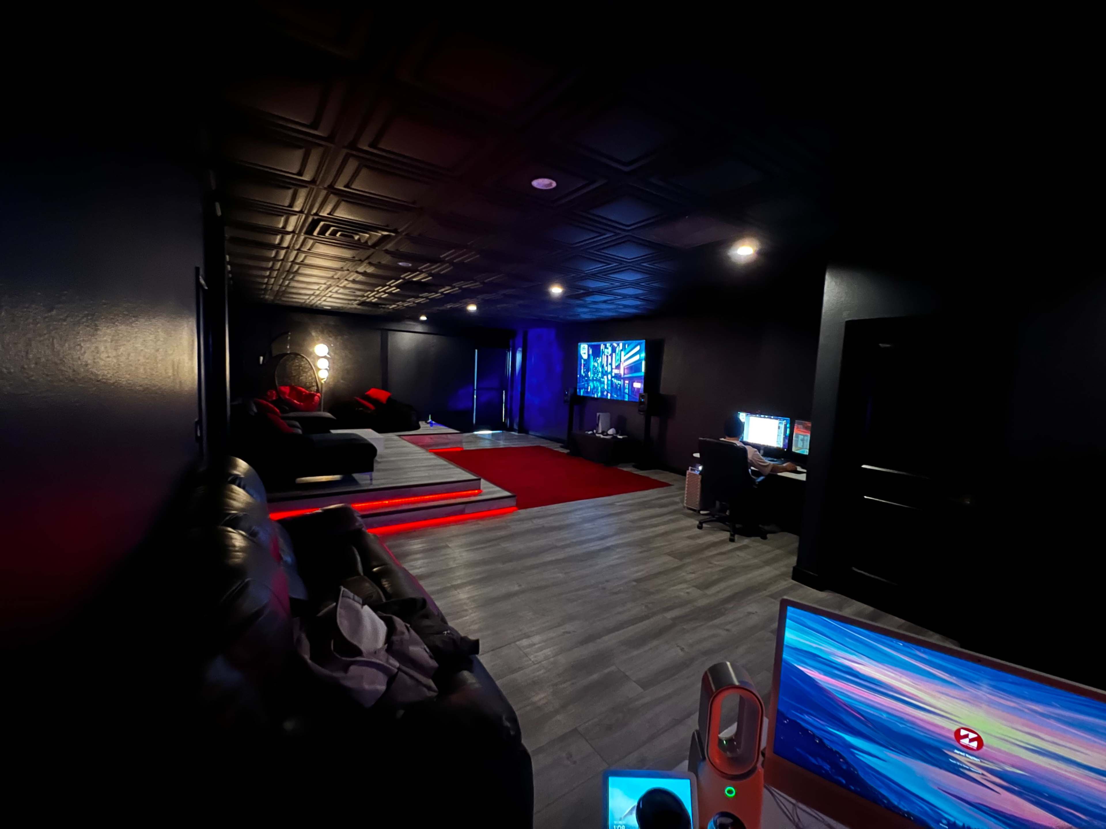 A dimly lit entertainment room features a large sectional sofa, a red carpet, a screen displaying colorful visuals, and two computer setups.