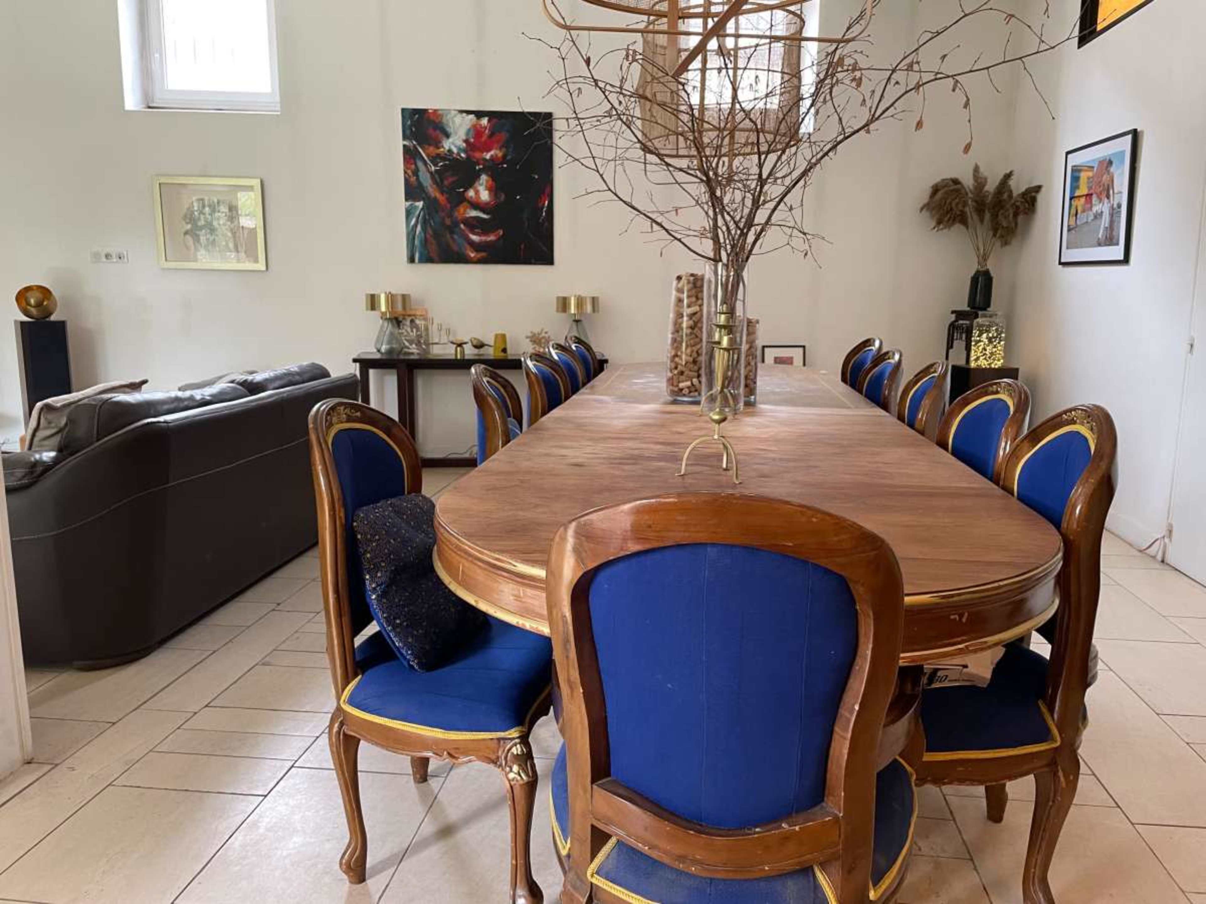 A long wooden dining table with blue upholstered chairs is set in a well-lit room featuring artwork on the walls.