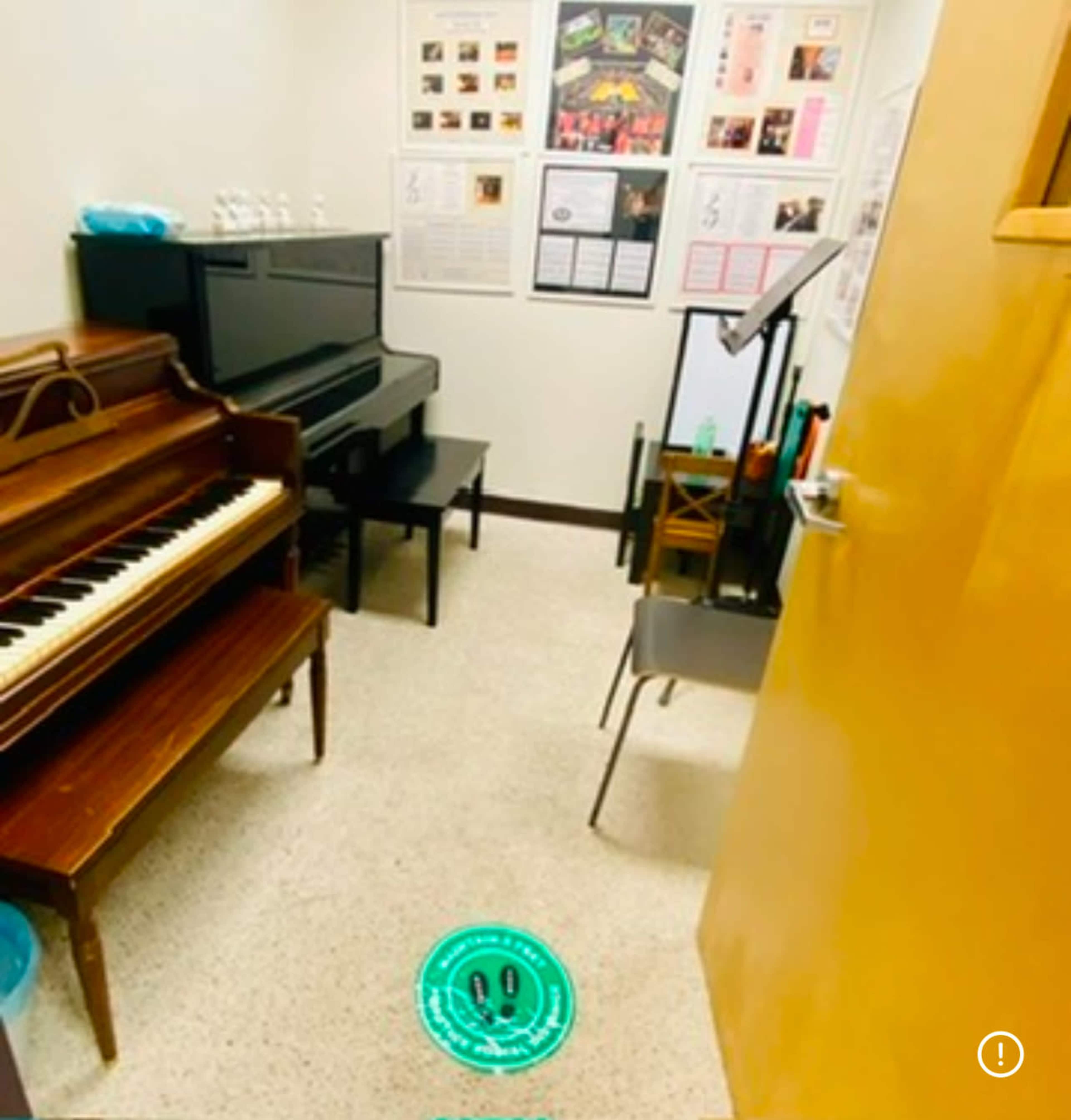 The image shows a small music practice room containing a grand piano, a black piano, a chair, a music stand, and various posters on the walls.