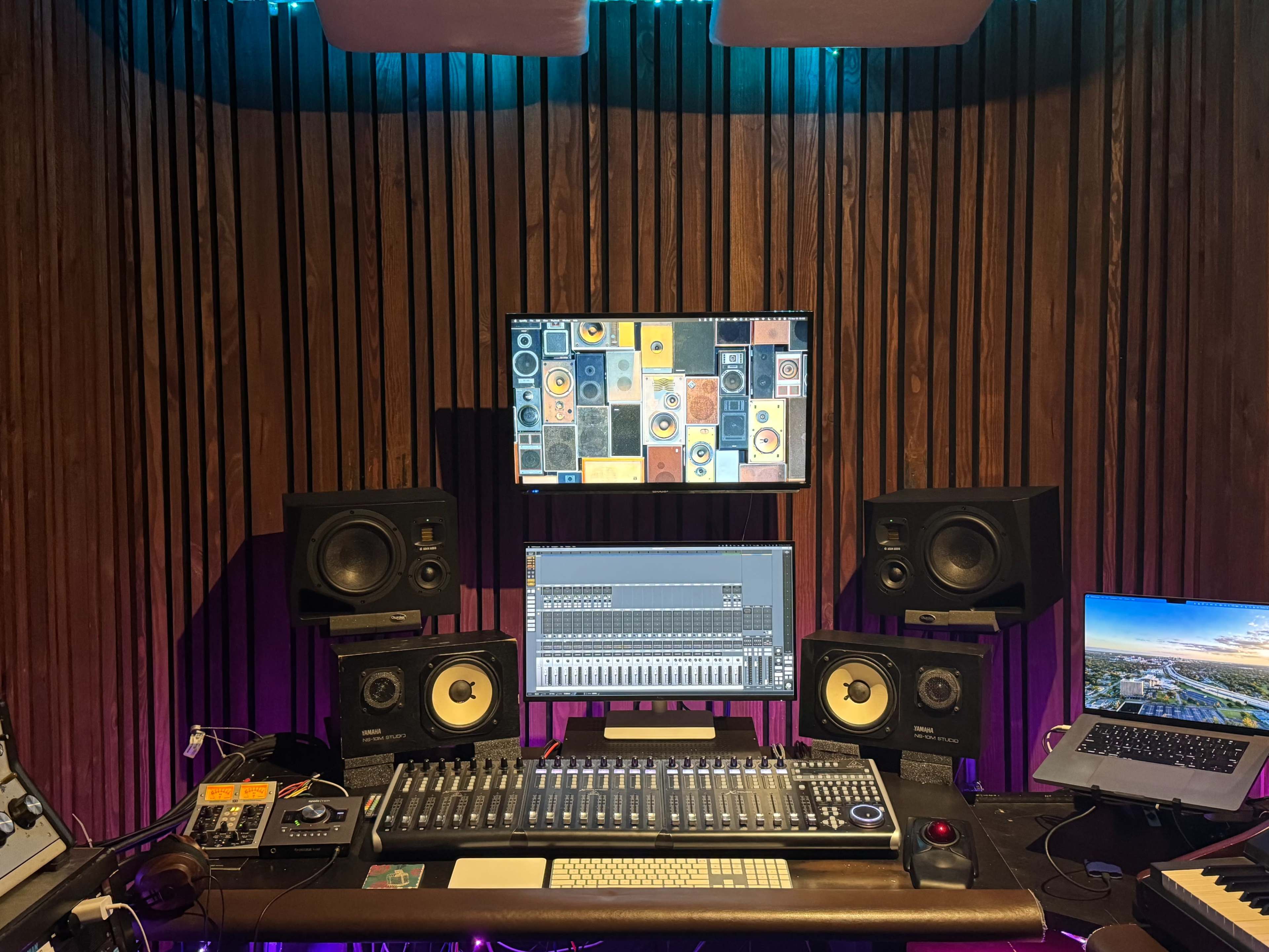 The image shows a music production studio with a mixing console, two studio monitors, a computer displaying audio software, and wooden-paneled walls.