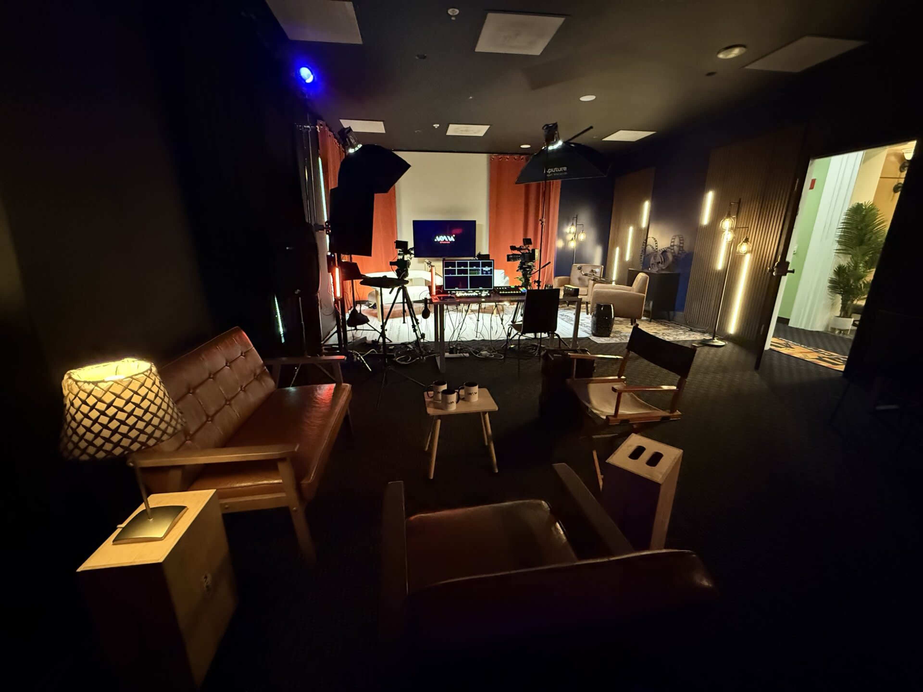 The image shows a darkly lit studio setup featuring several cameras and lighting equipment, with comfortable seating and decorative elements arranged around a central screen.