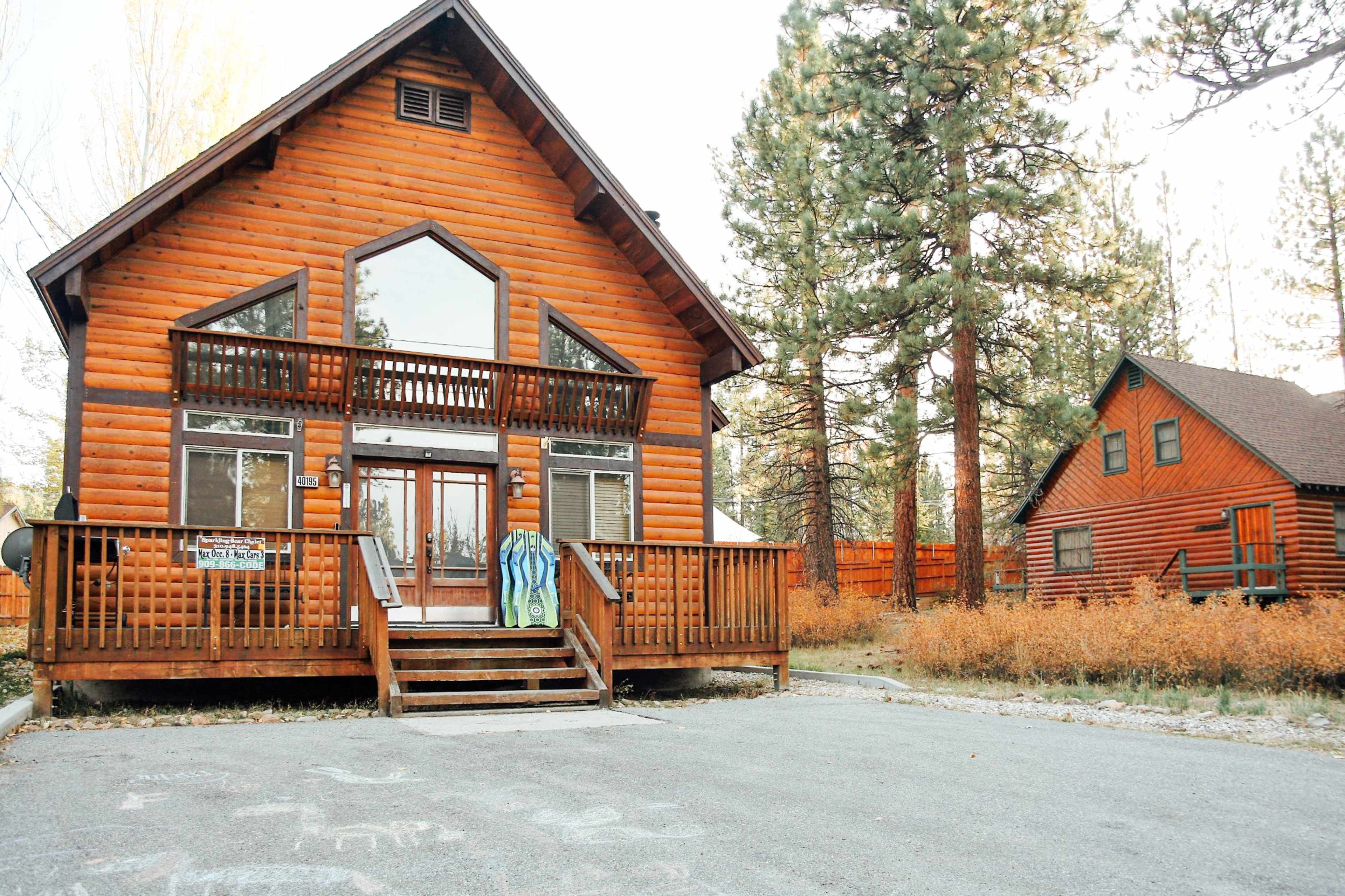 The image shows two log cabin-style houses surrounded by tall pine trees, with one larger cabin in the foreground and a smaller one in the background.
