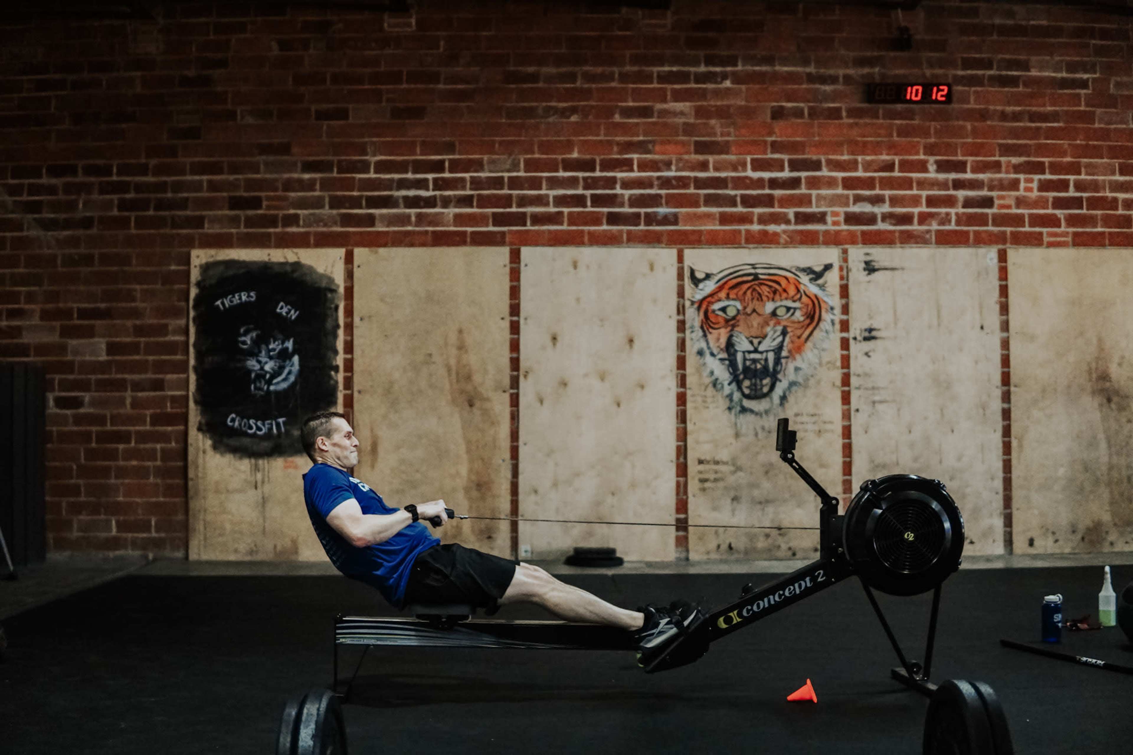 A man is exercising on a rowing machine in a gym with a brick wall and wooden panels.