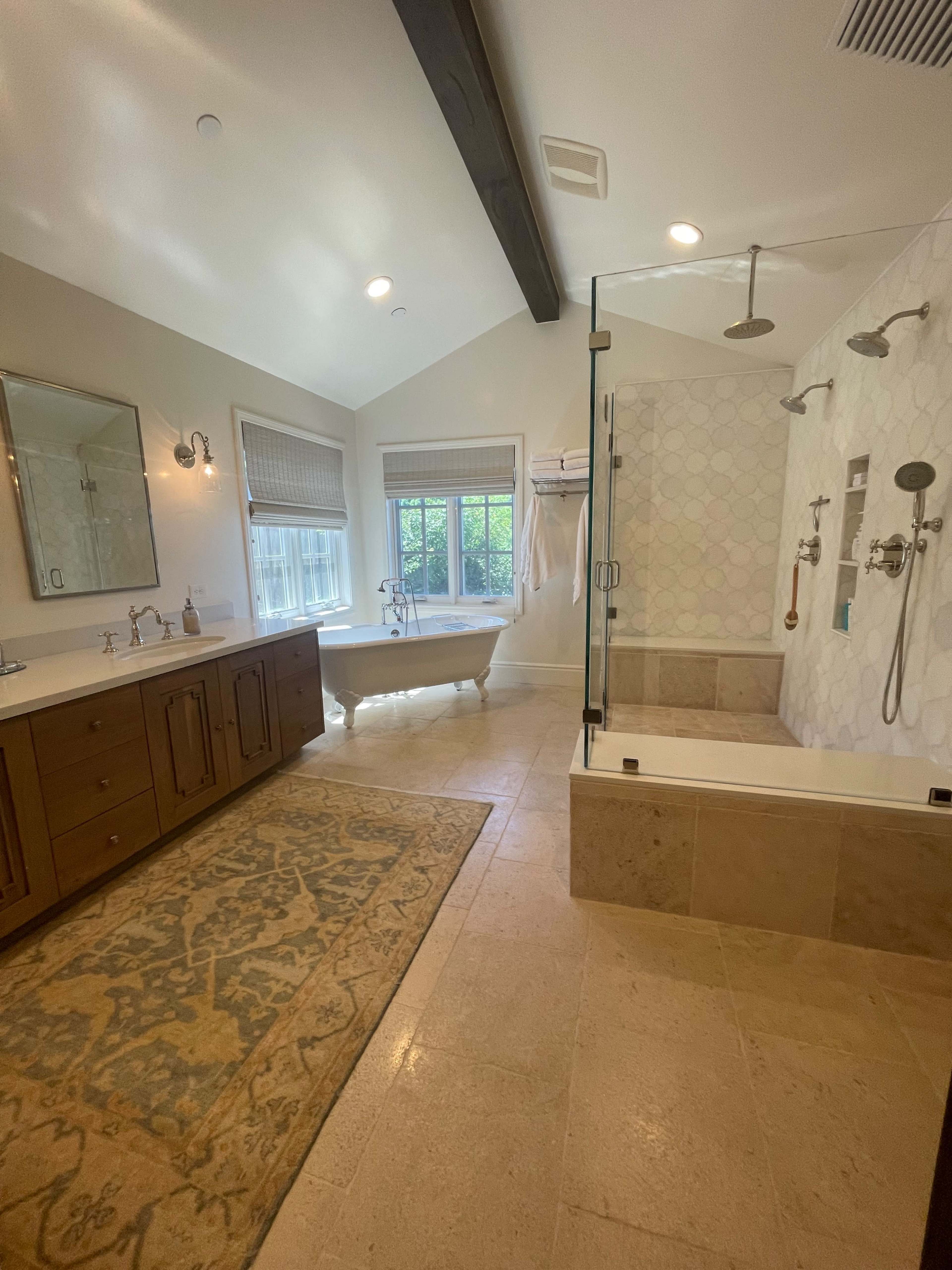 The bathroom features a freestanding bathtub, a double vanity, a glass-enclosed shower with multiple showerheads, and large windows providing natural light.