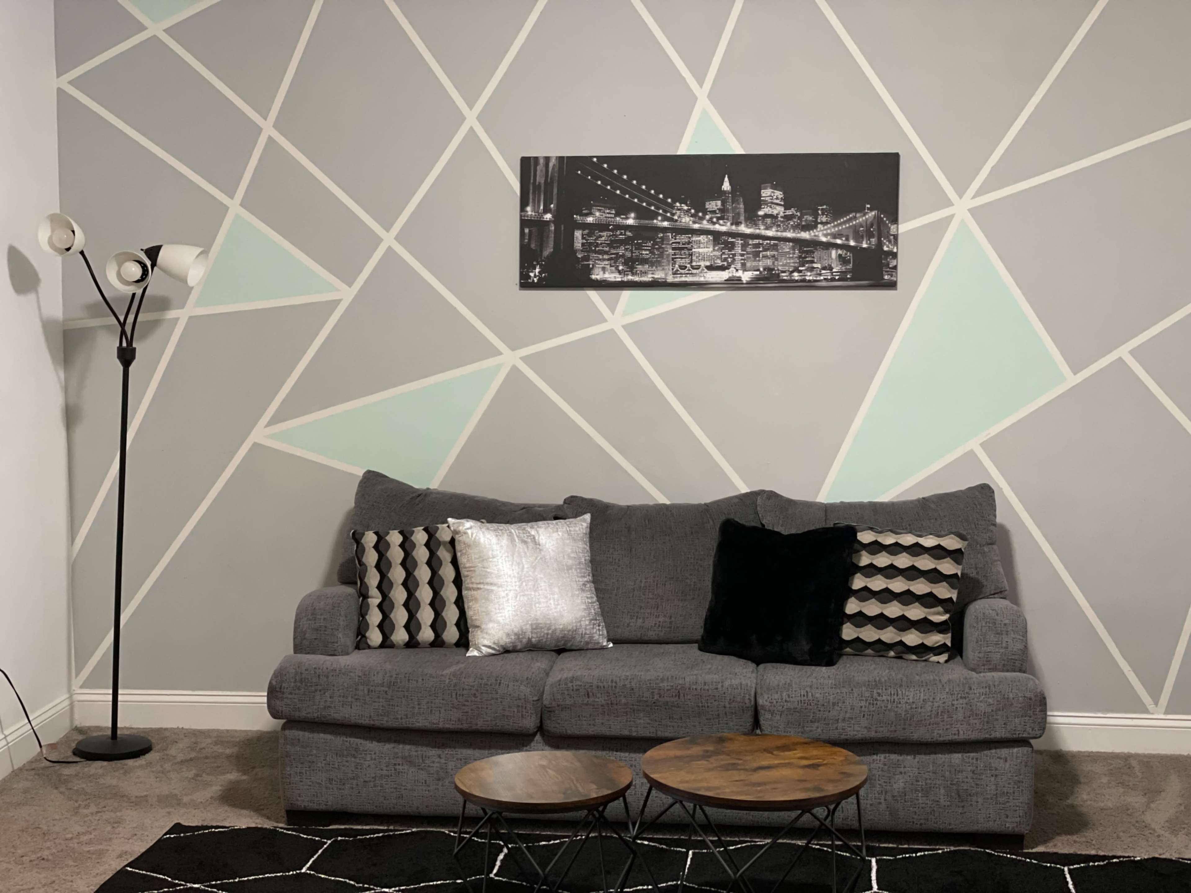 A gray couch with decorative pillows is placed against a wall featuring a geometric design, under a black-and-white photograph of a city skyline, with two round coffee tables in front.
