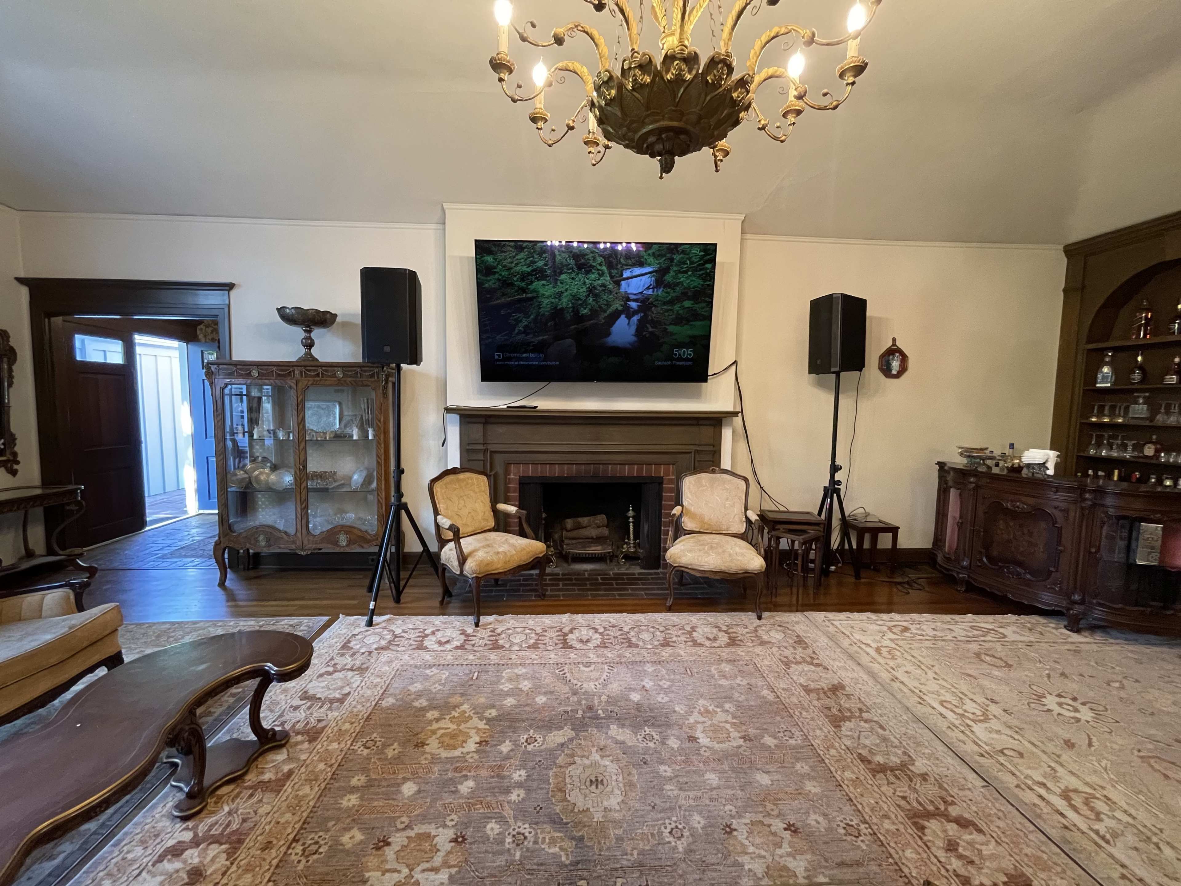 The room features a large flat-screen television mounted above a fireplace, flanked by two speakers, with vintage furniture and a chandelier overhead.
