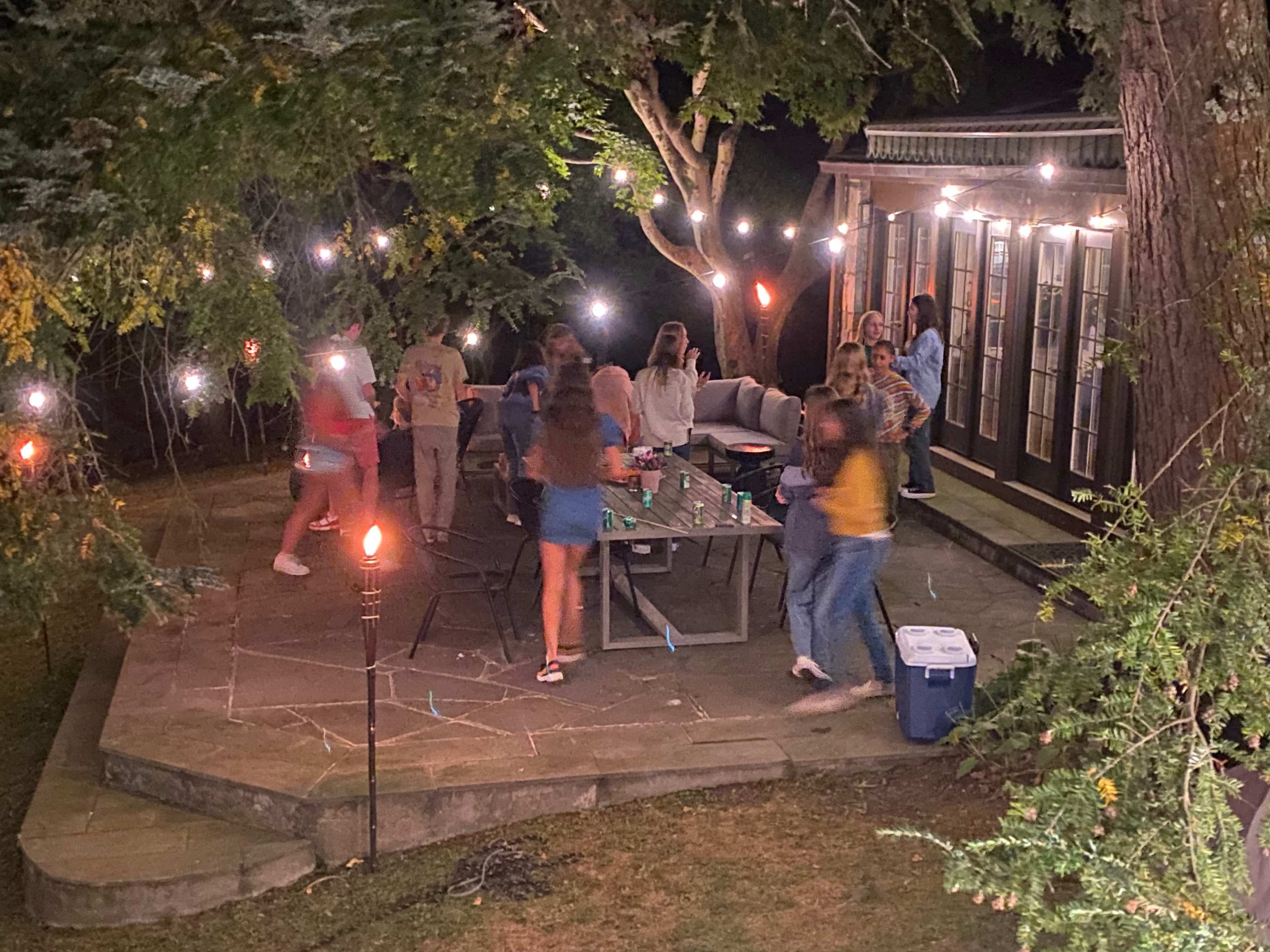 A group of people gather around a table on a stone patio, surrounded by string lights and trees in a backyard setting at night.