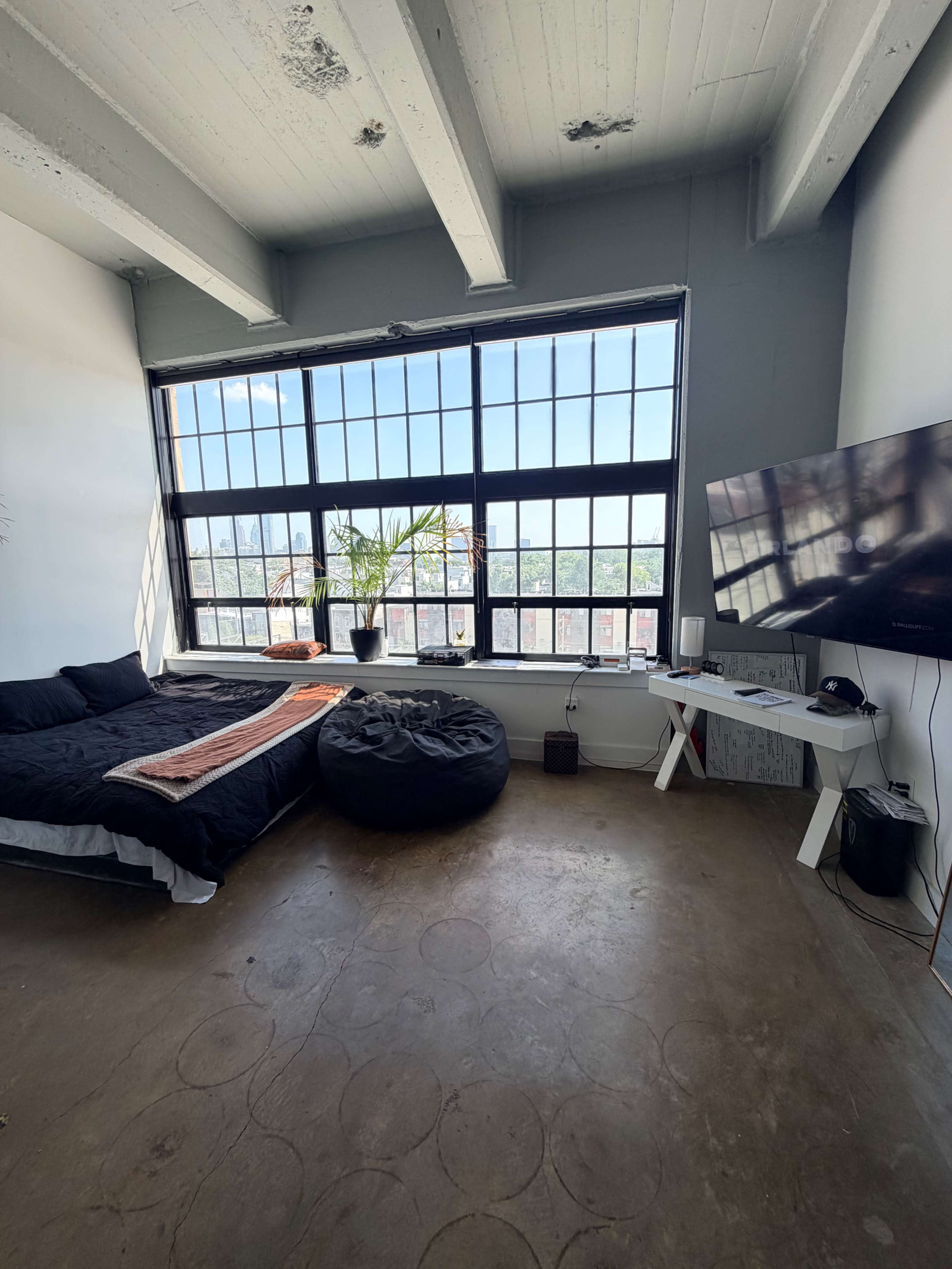 The image shows a minimalist bedroom with a large window, a bed covered in black bedding, a bean bag chair, and a television mounted on a white stand.