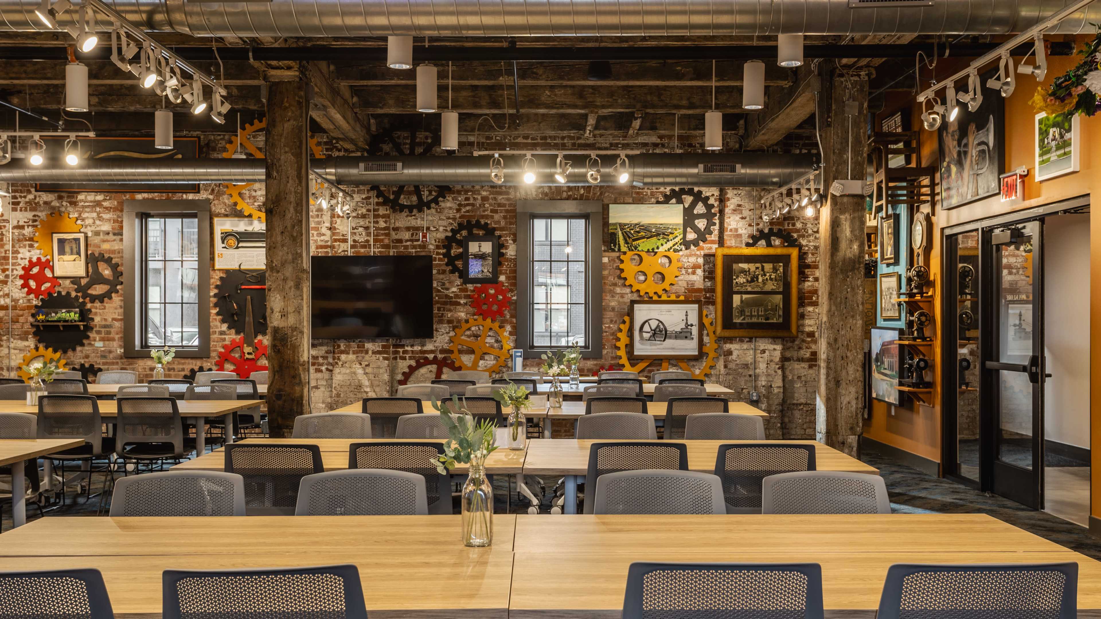 The image shows a spacious indoor workspace with modern tables and chairs, exposed brick walls adorned with colorful gears and framed photographs, and ample natural light coming through large windows.