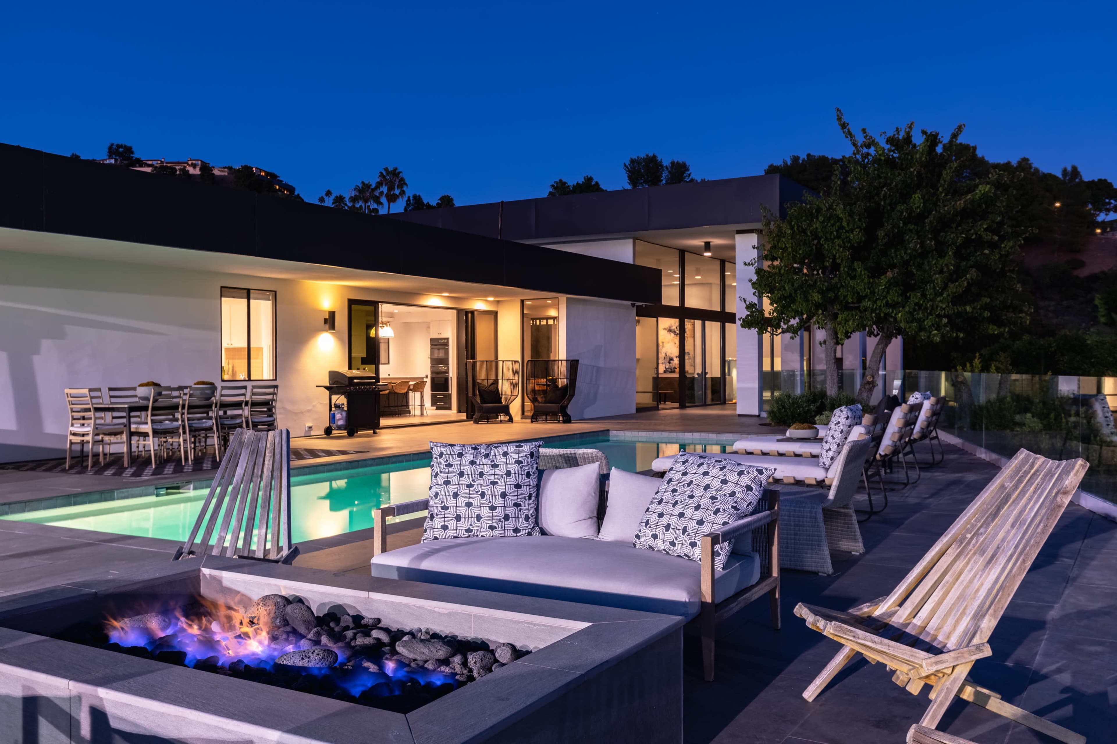 A modern backyard features a swimming pool, cozy seating around a fire pit, and a stylish house in the background, illuminated at night.