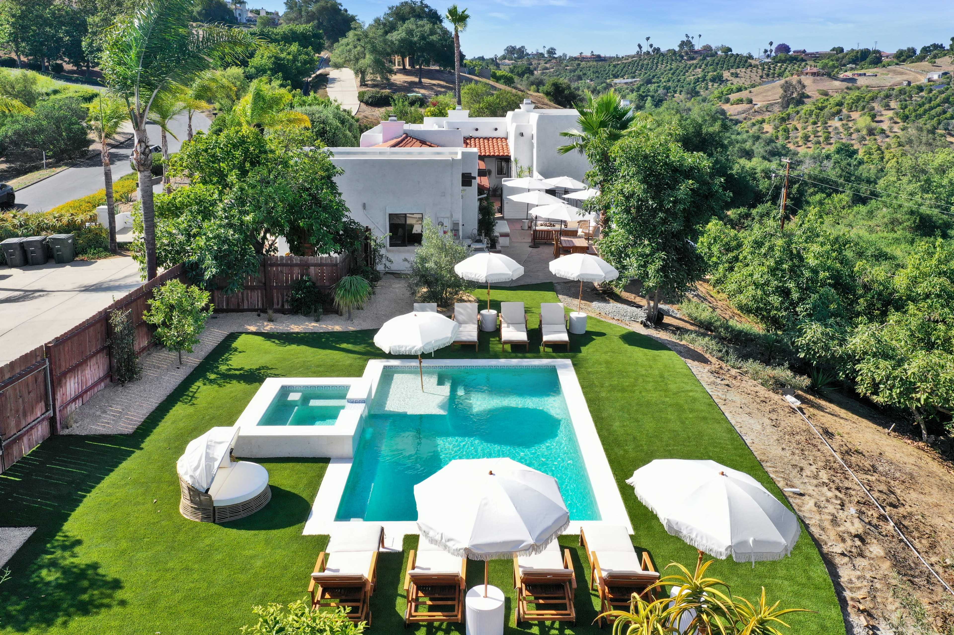The image shows a modern backyard with a swimming pool, spa, lounge chairs, and white umbrellas surrounded by greenery and a hillside view.