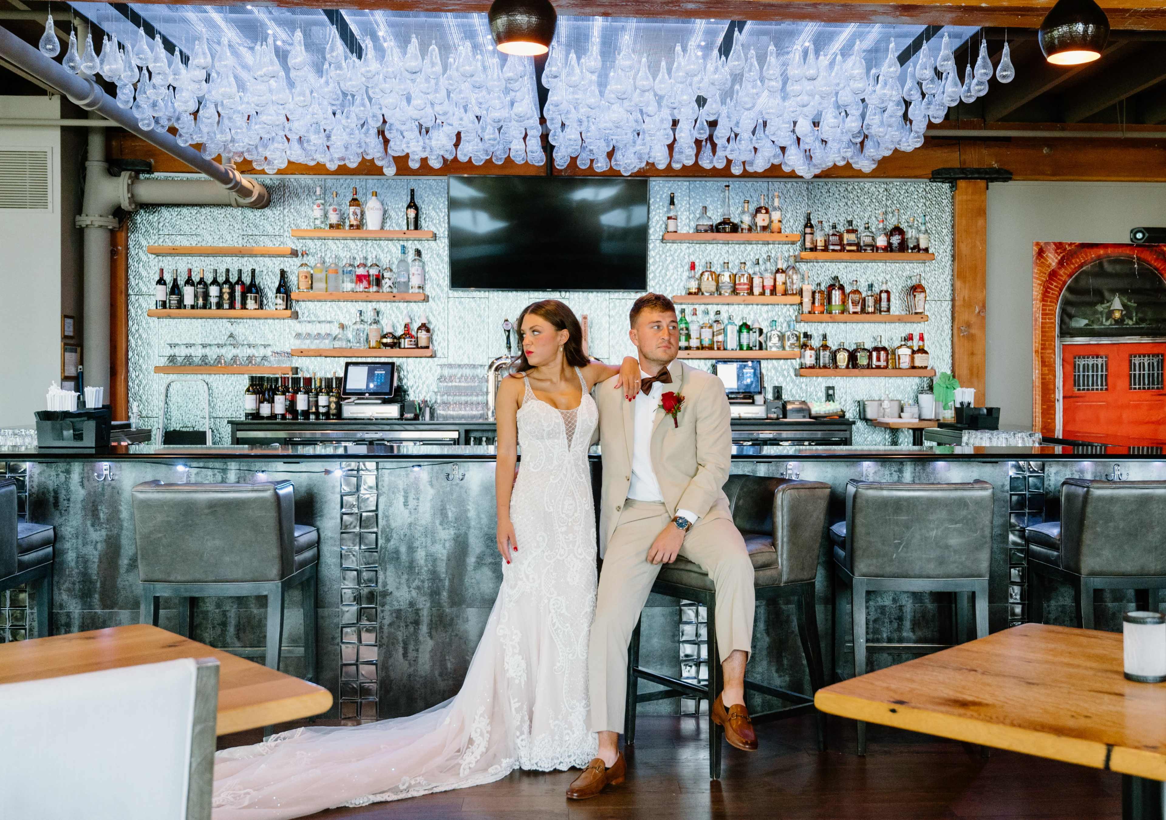 A bride in a lace wedding dress stands beside a groom in a beige suit at a bar with an array of bottles displayed on shelves behind them.