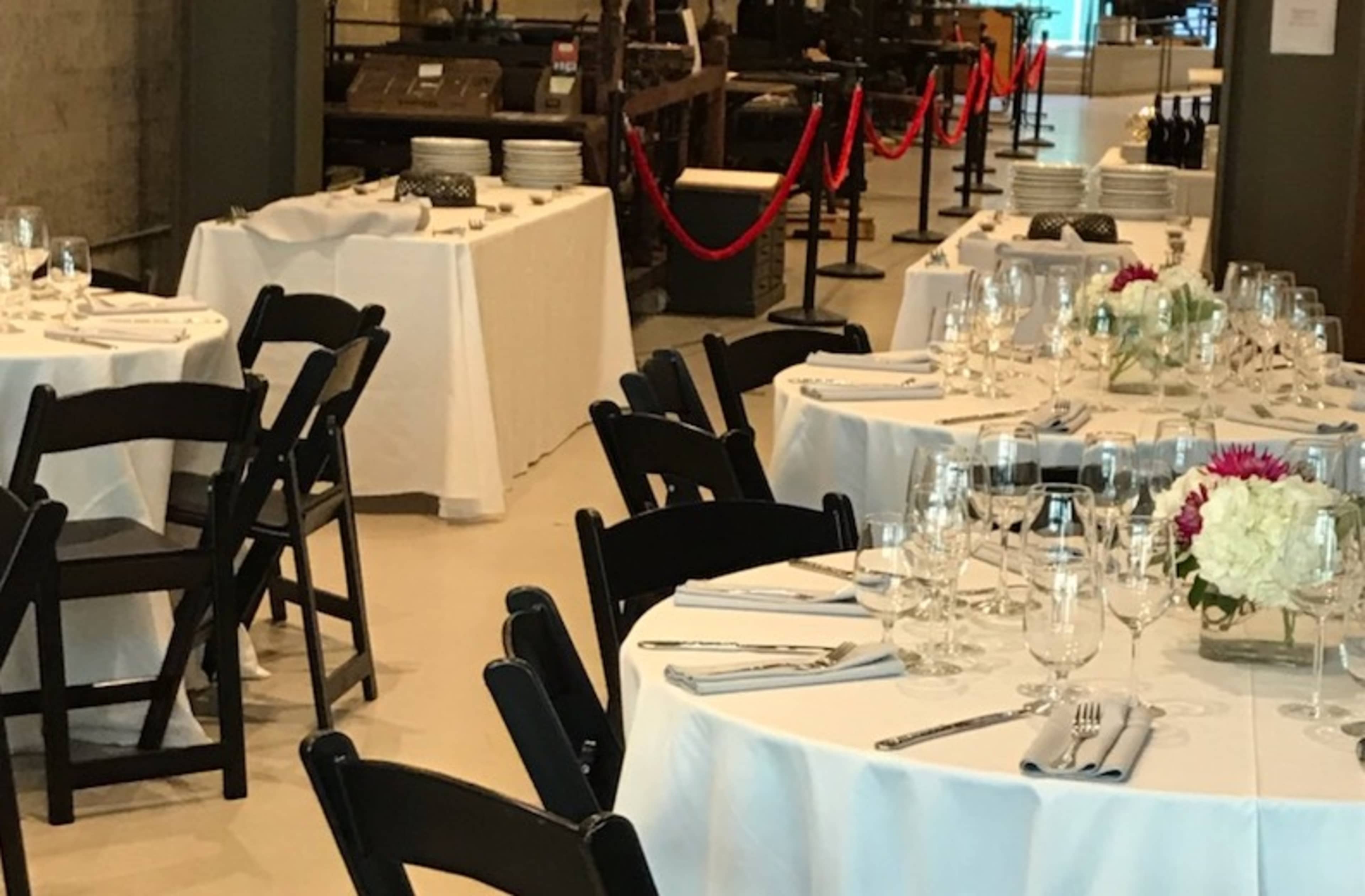 A dining area with neatly arranged tables covered in white tablecloths and set with glassware and utensils, adjacent to a buffet table with plates and a red rope barrier.