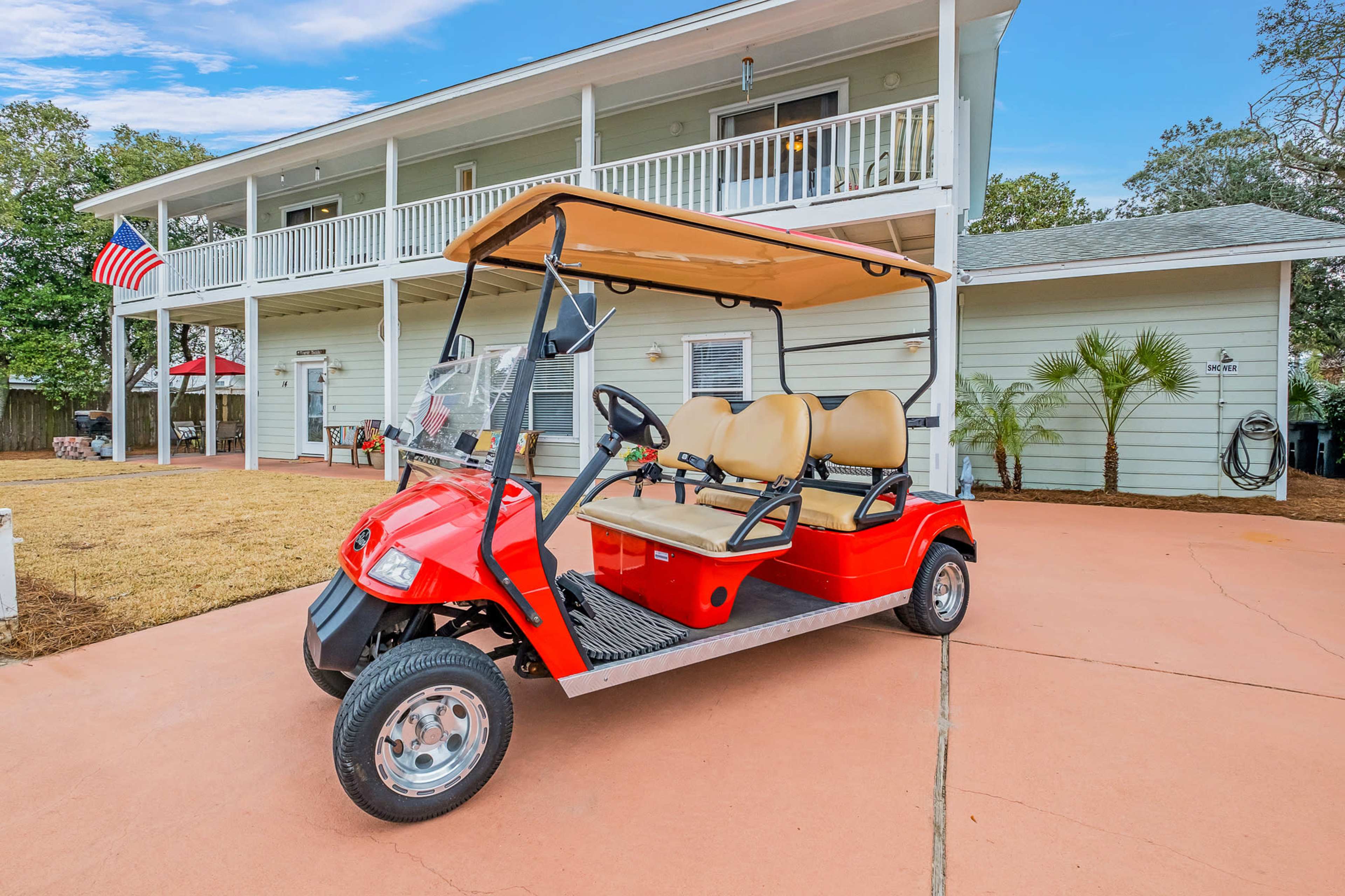 A red golf cart is parked on a concrete driveway in front of a two-story house with a porch and an American flag.