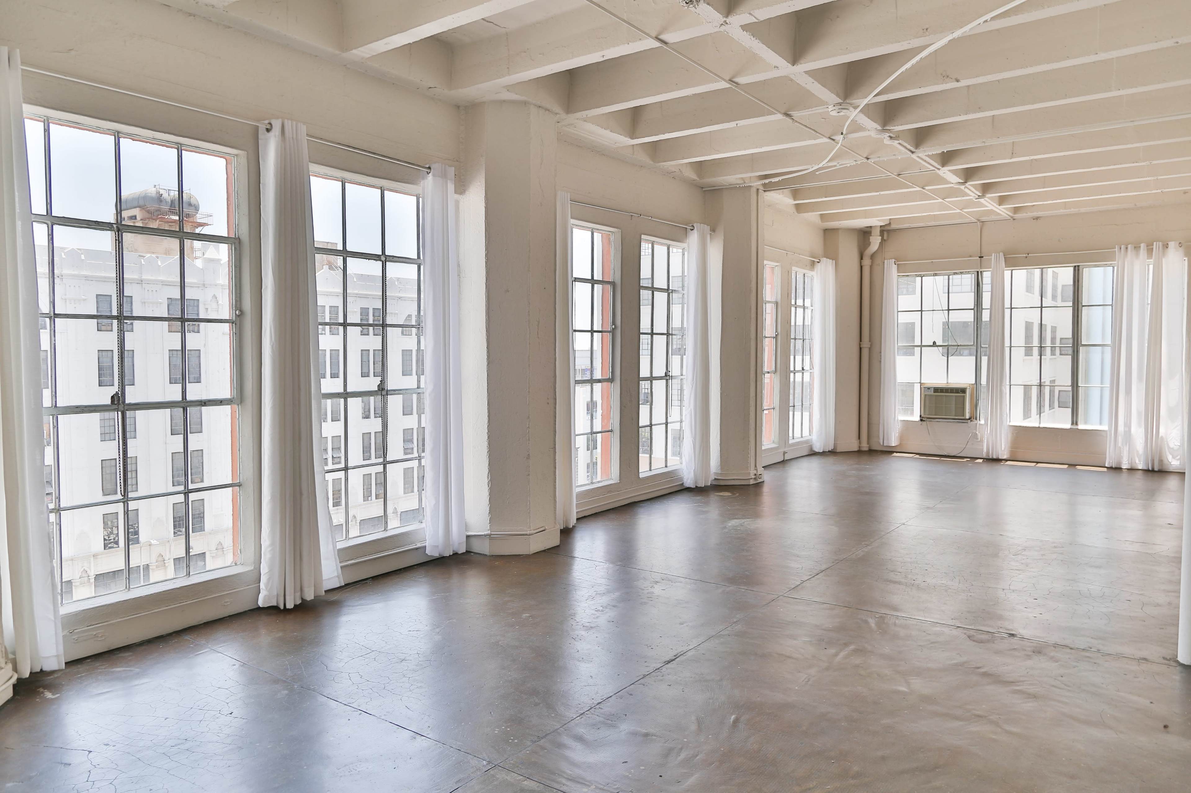 A spacious, empty room with large windows and polished concrete floors, featuring white walls and light curtains.
