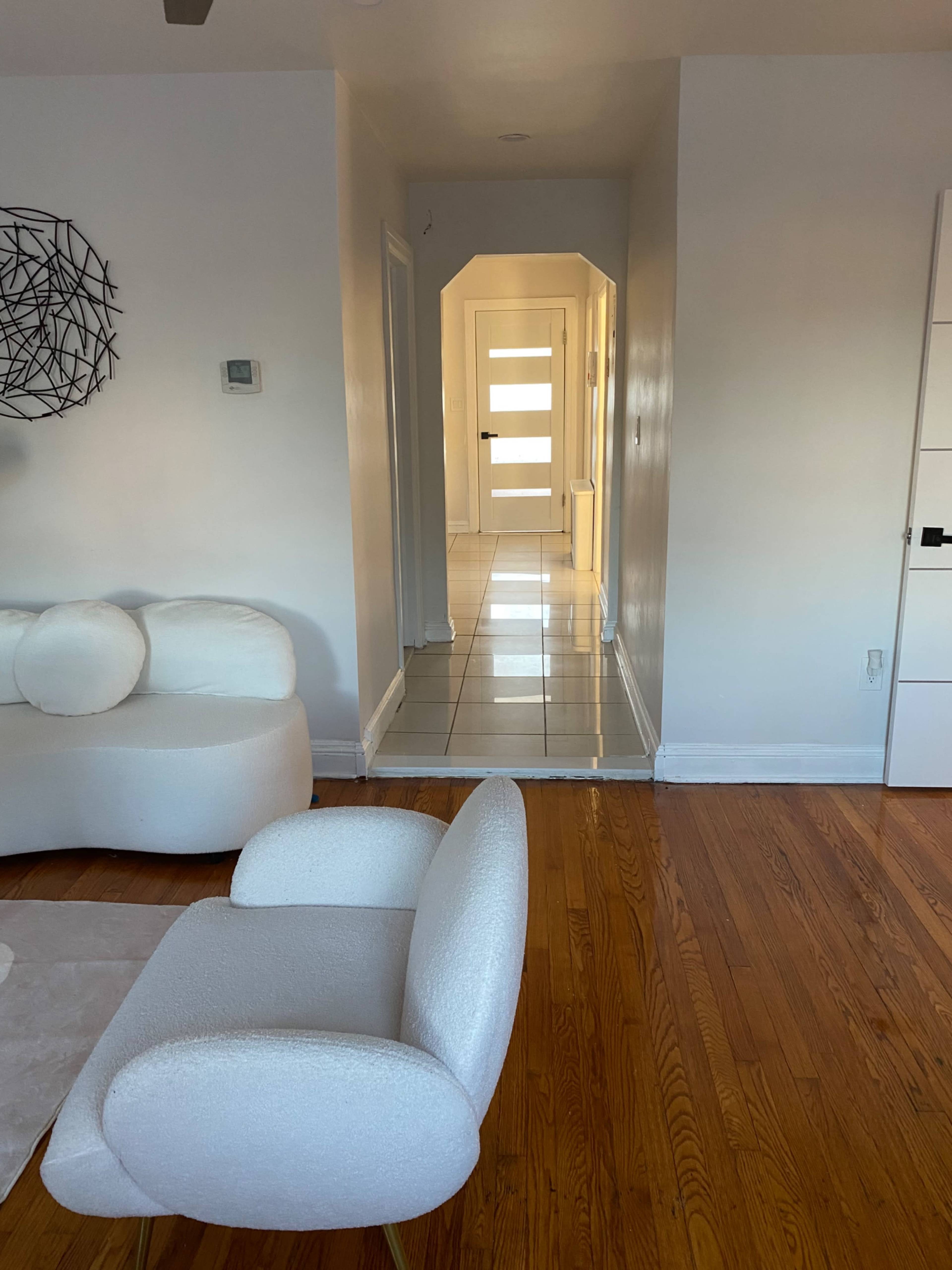 A white modern living room features a plush chair and a hallway leading to a door with frosted glass panels.