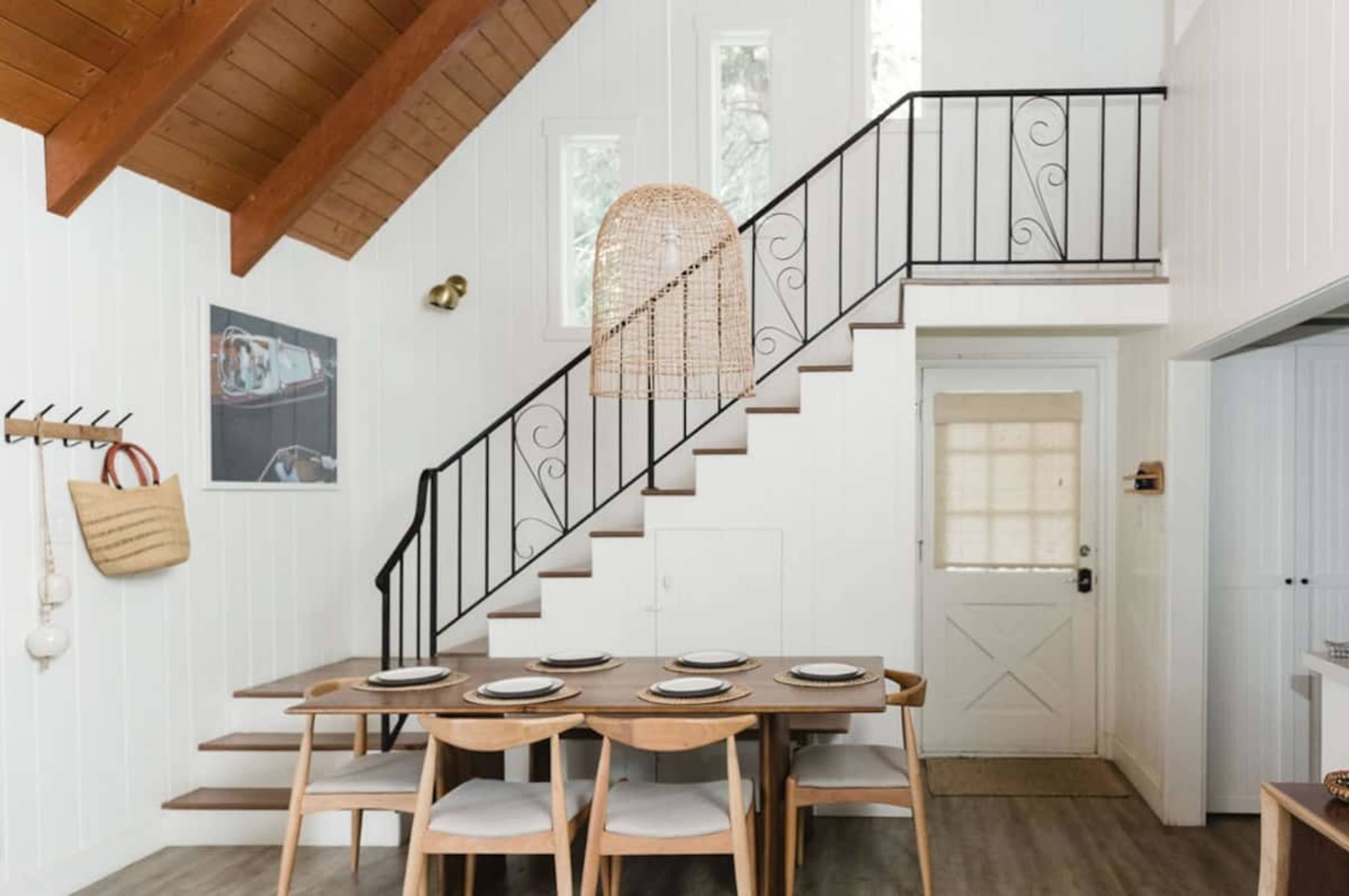 A spacious interior features a dining table set for six, a staircase with a decorative railing, and large windows allowing natural light.