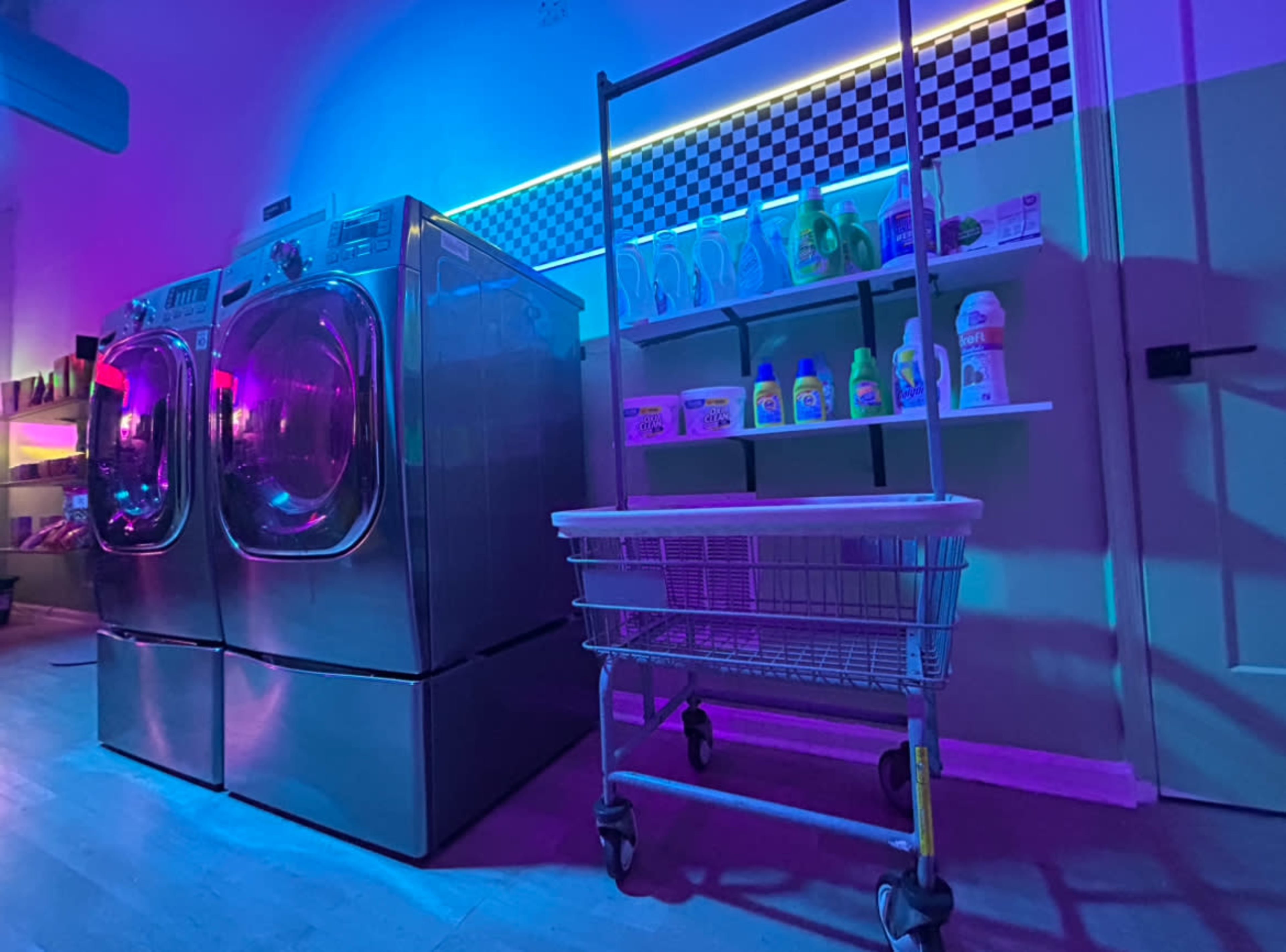 The image shows a laundry room with two silver washing machines, a white laundry cart, and shelves stocked with various detergent products, all illuminated by colorful lighting.