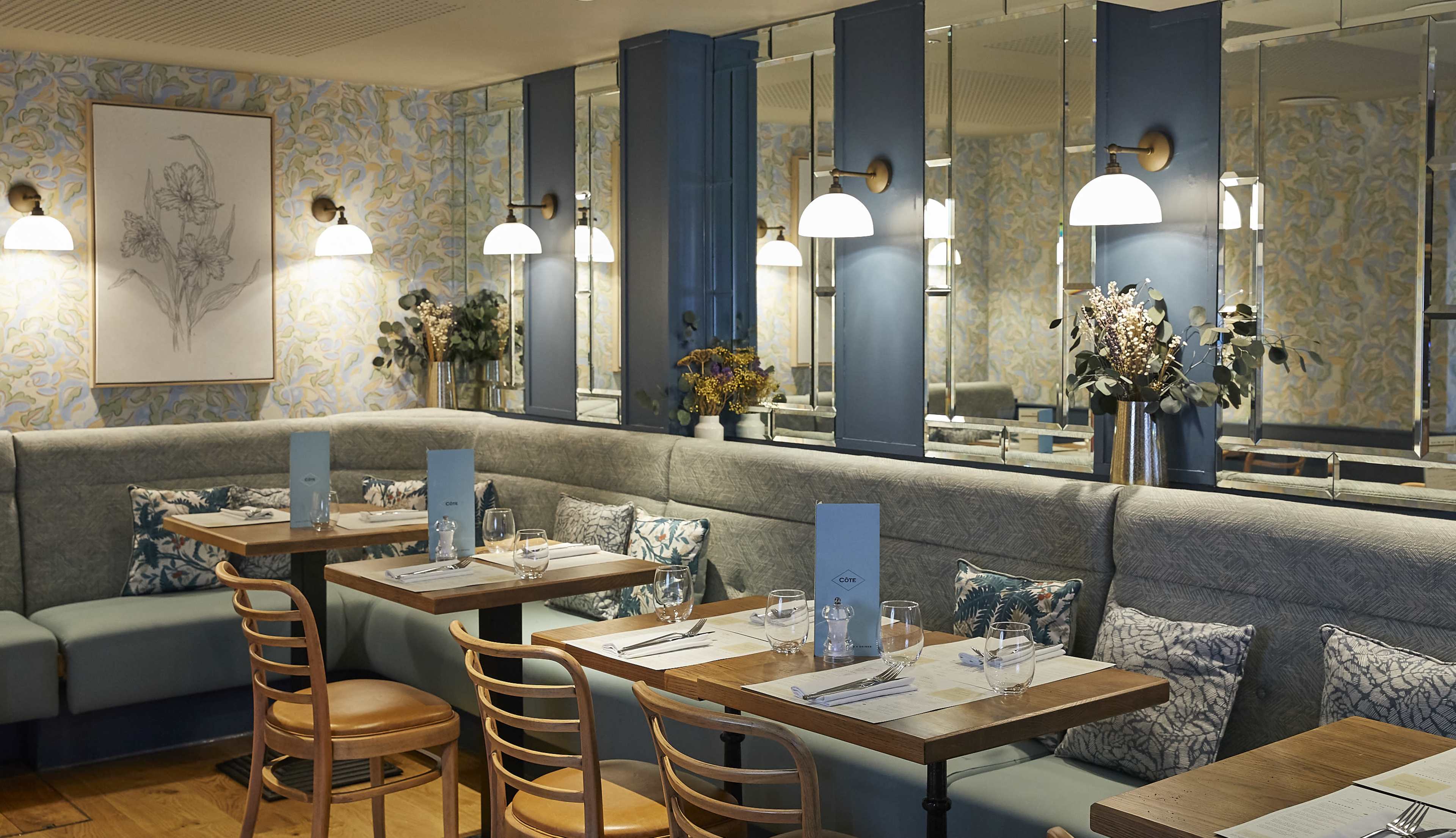 The image shows a restaurant interior with blue and beige decor, featuring wooden tables, upholstered benches, and floral accents.