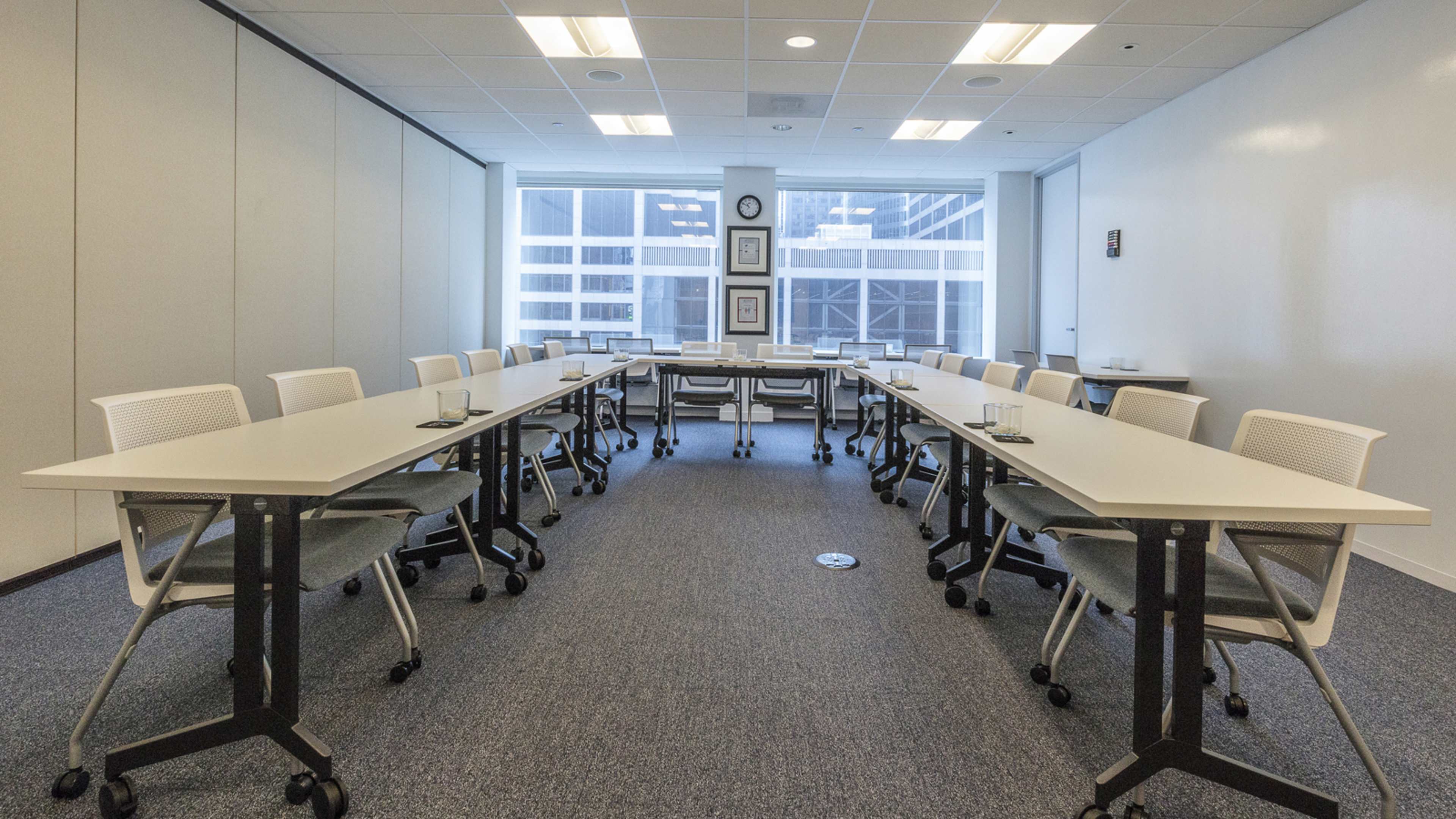 A modern conference room features two long tables arranged in a U-shape, surrounded by white chairs, with large windows allowing natural light to fill the space.