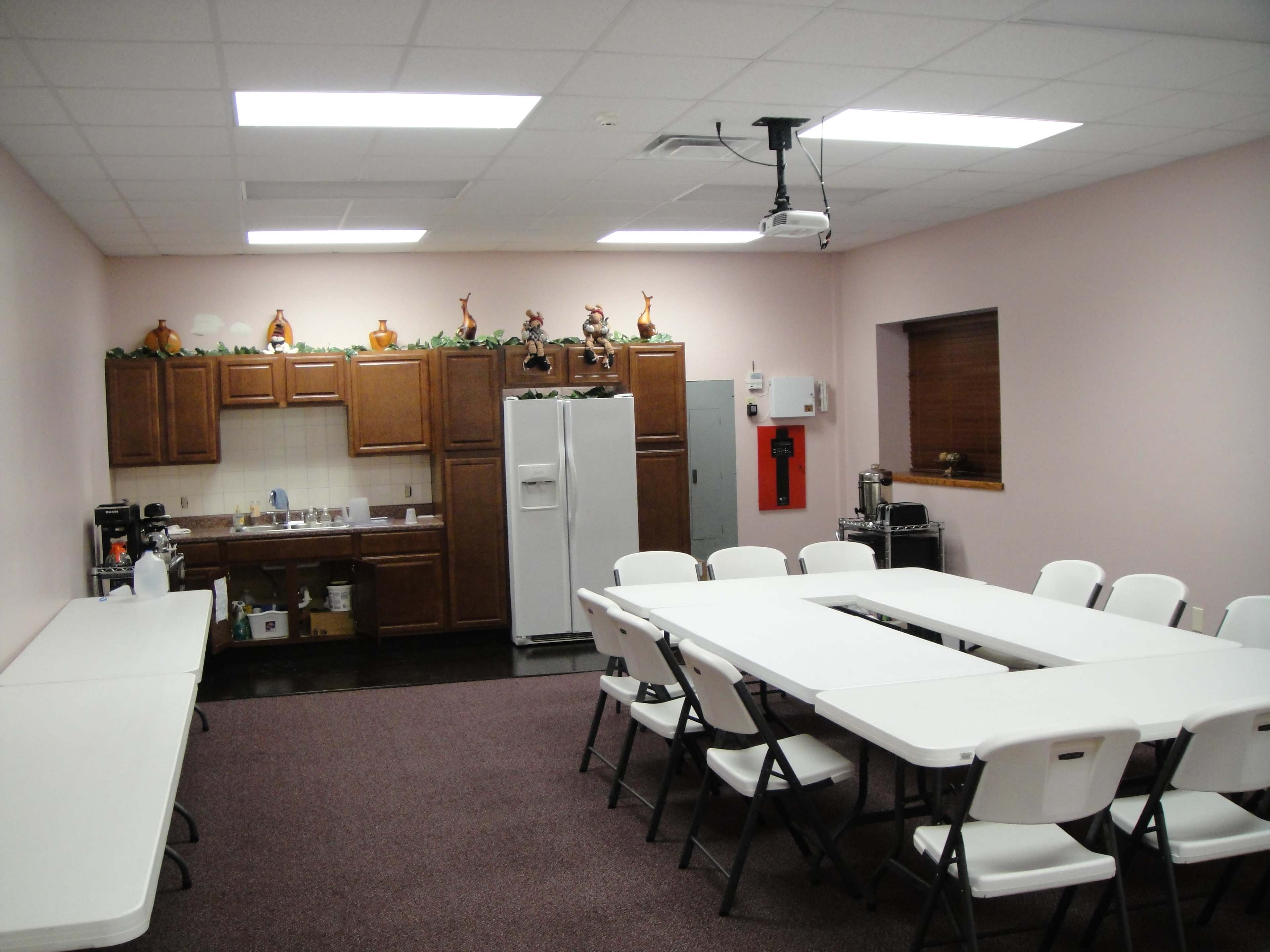A meeting room with a long table, chairs, a kitchenette area, and a wall-mounted projector.