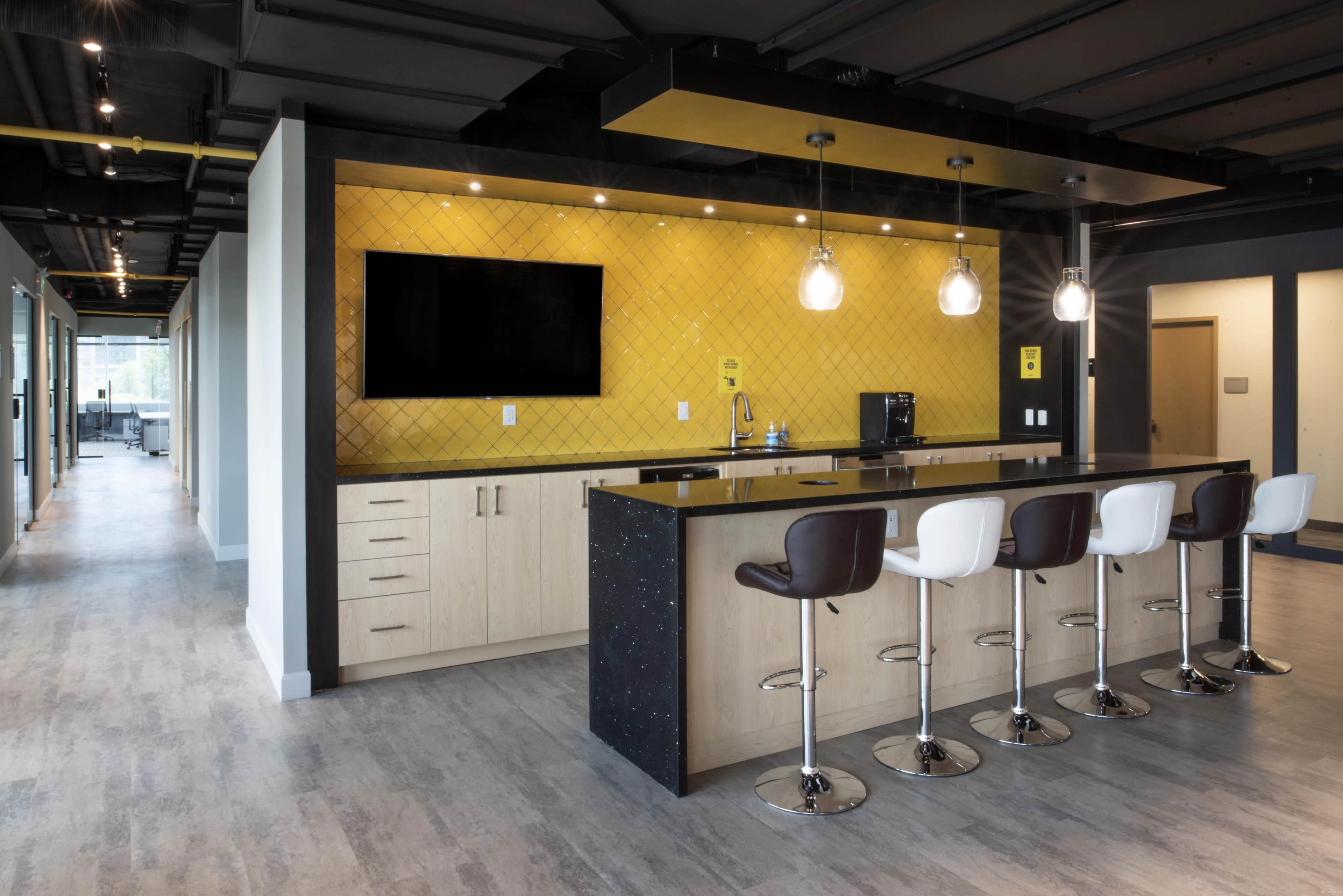 A modern kitchen area in an office with a long bar counter, four high stools, a television mounted on the wall, and yellow tiles as a backdrop.
