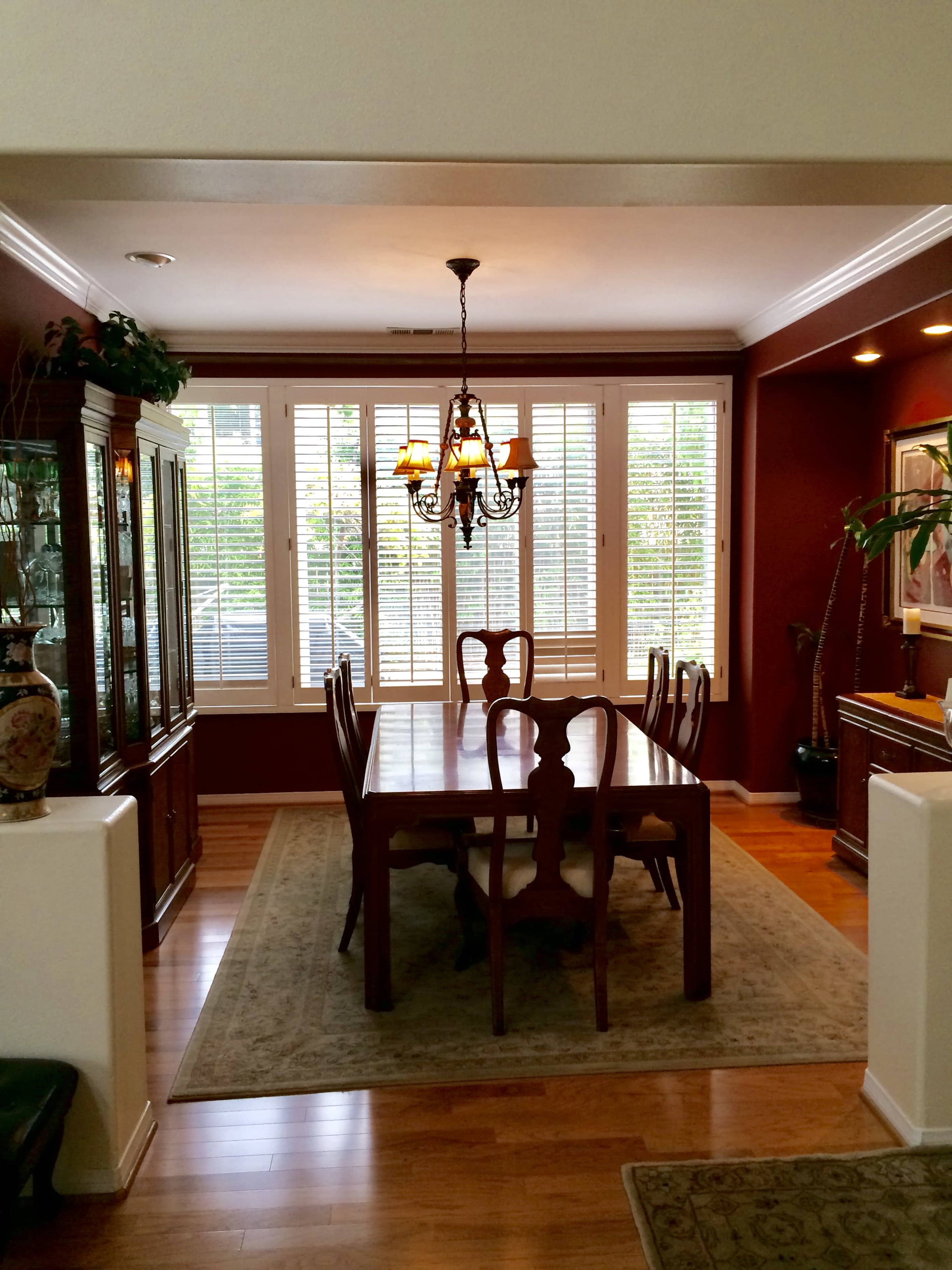 A dining room features a large wooden table surrounded by chairs, with a chandelier overhead and windows dressed in white shutters.