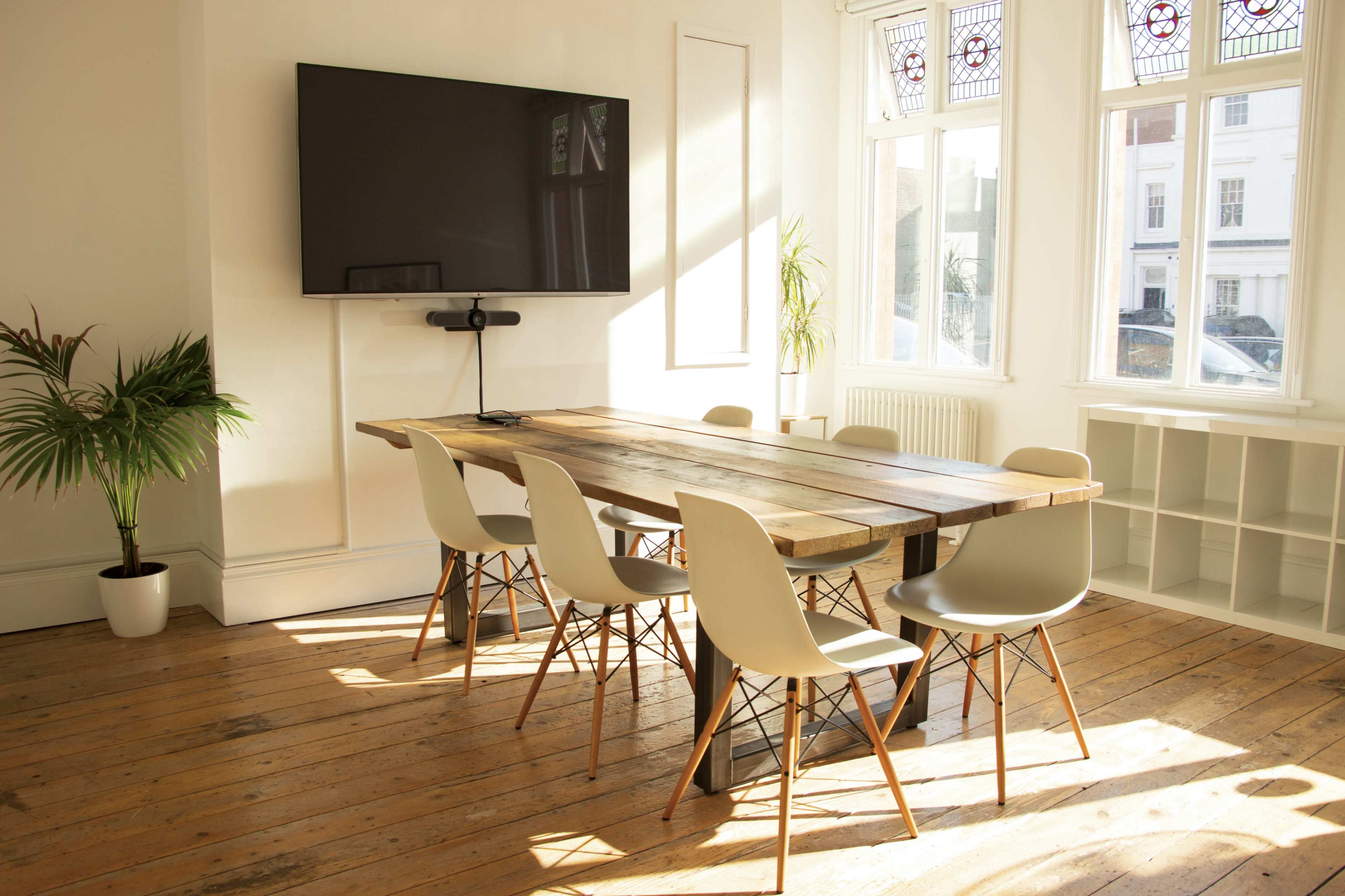 A bright, modern meeting room features a long wooden table surrounded by six chairs, with a large wall-mounted screen and a potted plant in the corner.