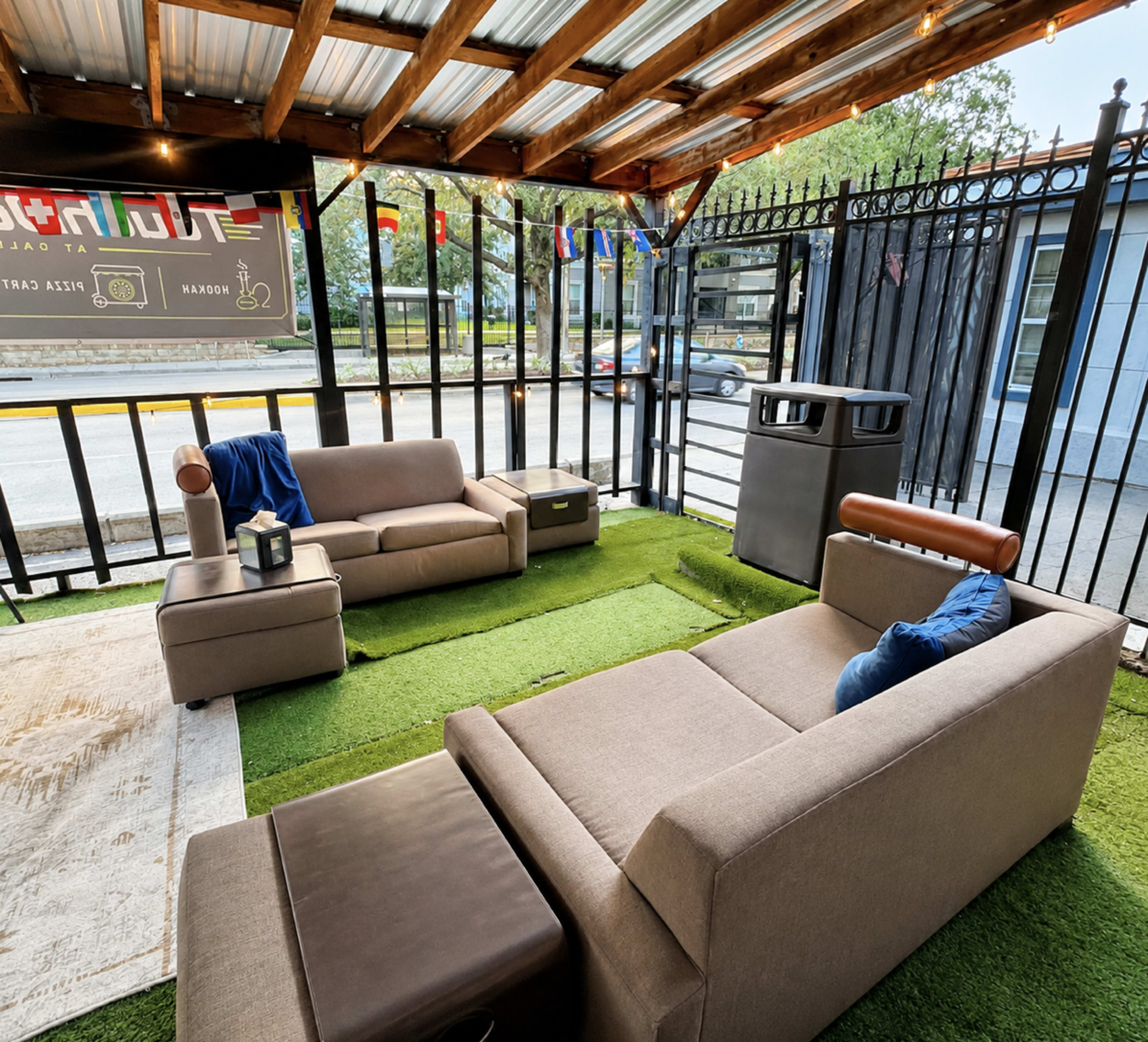 The image shows a cozy outdoor seating area with two modern sofas arranged on artificial grass, surrounded by a railing and decorative flags.