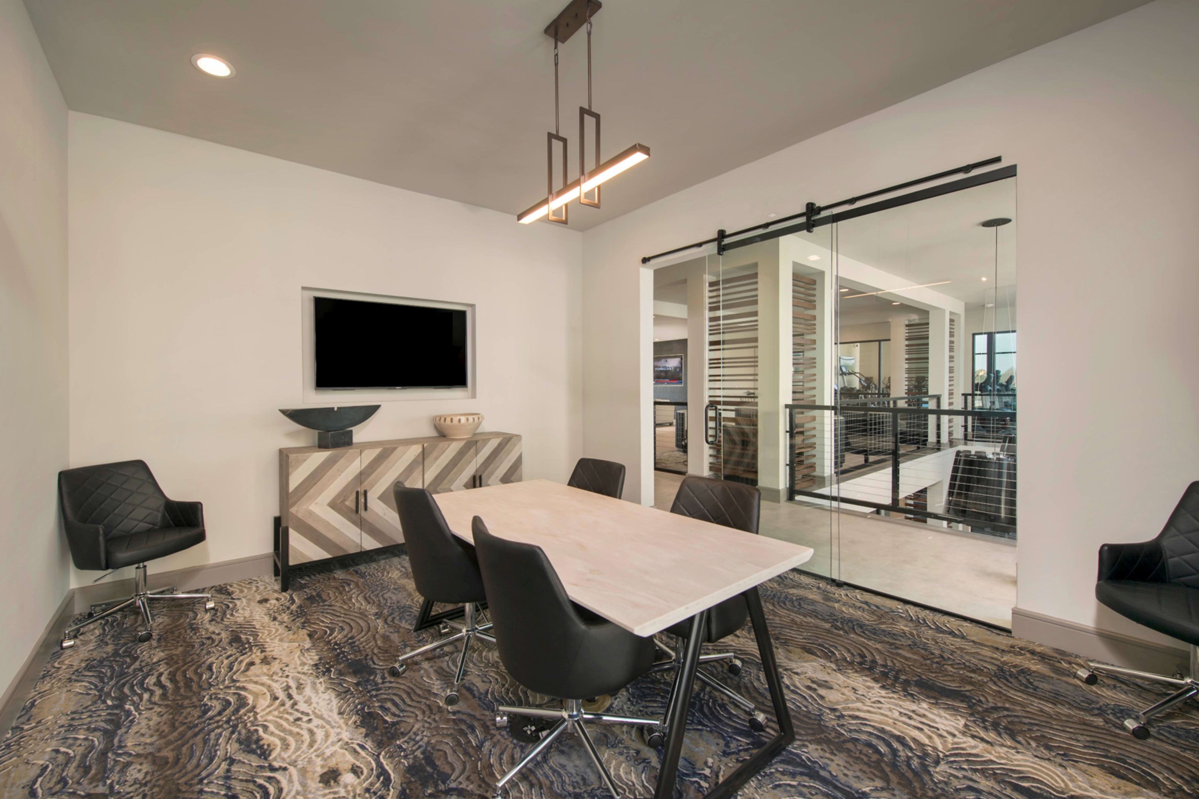 A modern conference room features a rectangular table surrounded by black chairs, with a television mounted on the wall and large glass doors leading to an adjacent space.