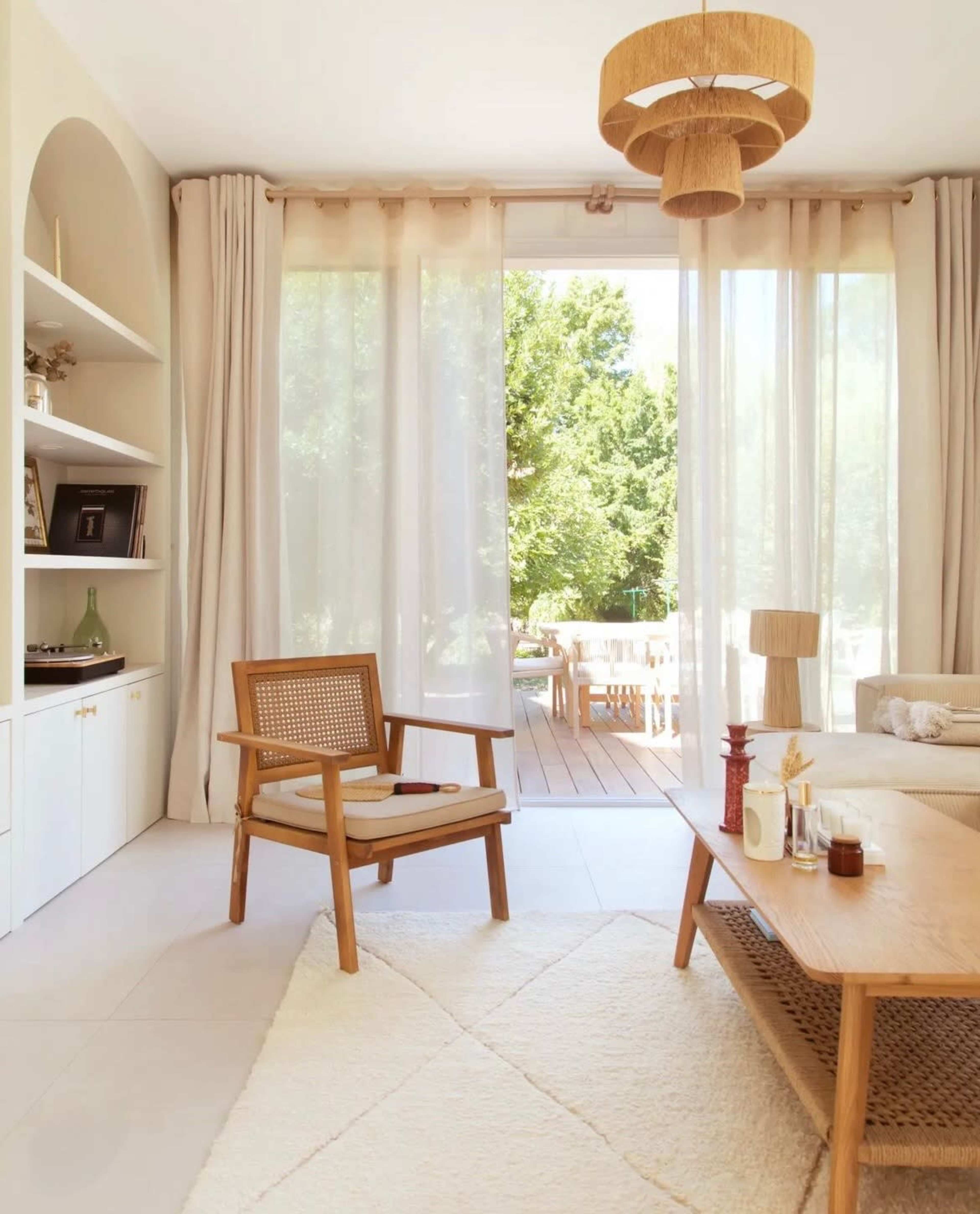 A cozy living room features a wooden armchair, a light wooden coffee table, and large windows displaying an outdoor deck surrounded by greenery.
