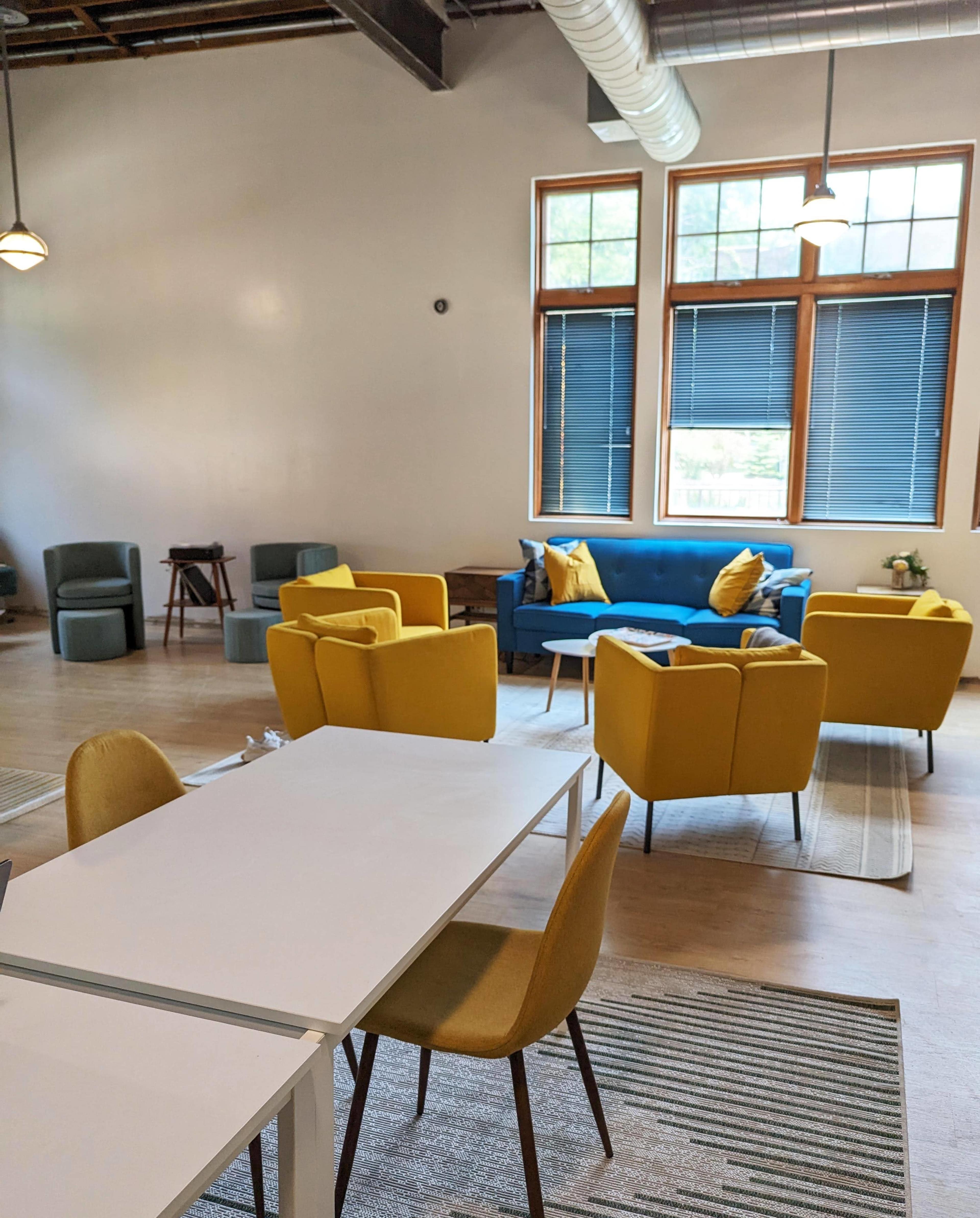 The image shows a modern, open space with a mix of seating areas, including yellow and blue chairs, a white table, and large windows letting in natural light.