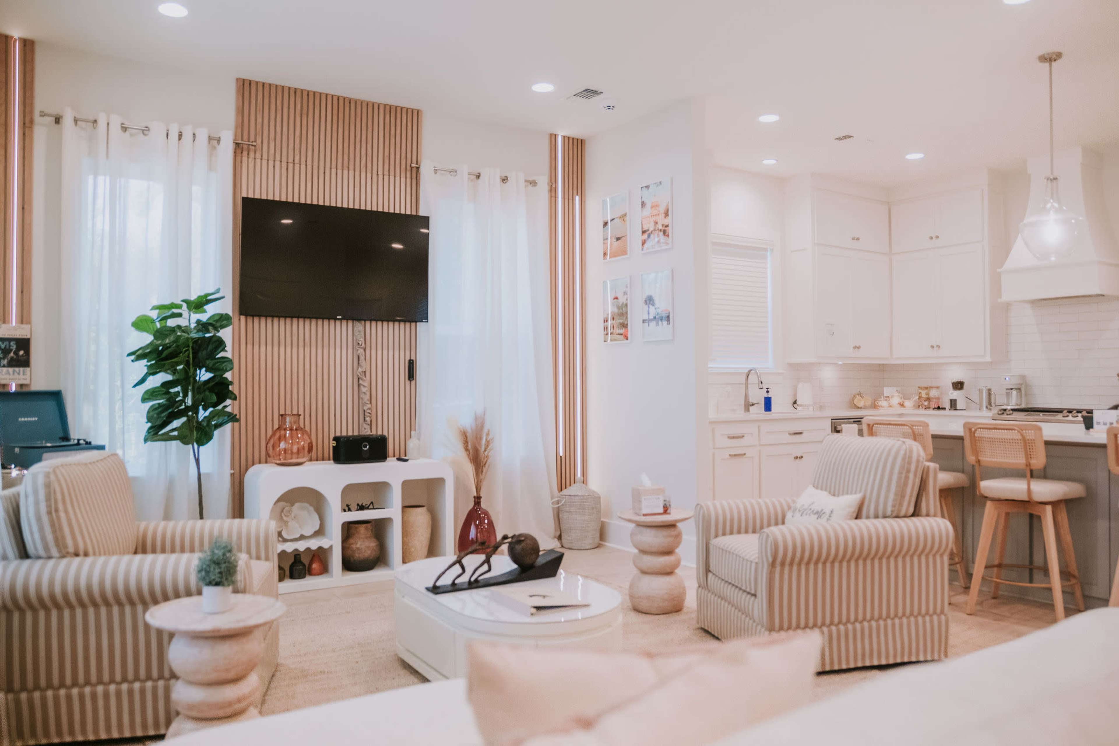 The image shows a modern living room with light-colored furniture, a wall-mounted television, and a kitchen area visible in the background.