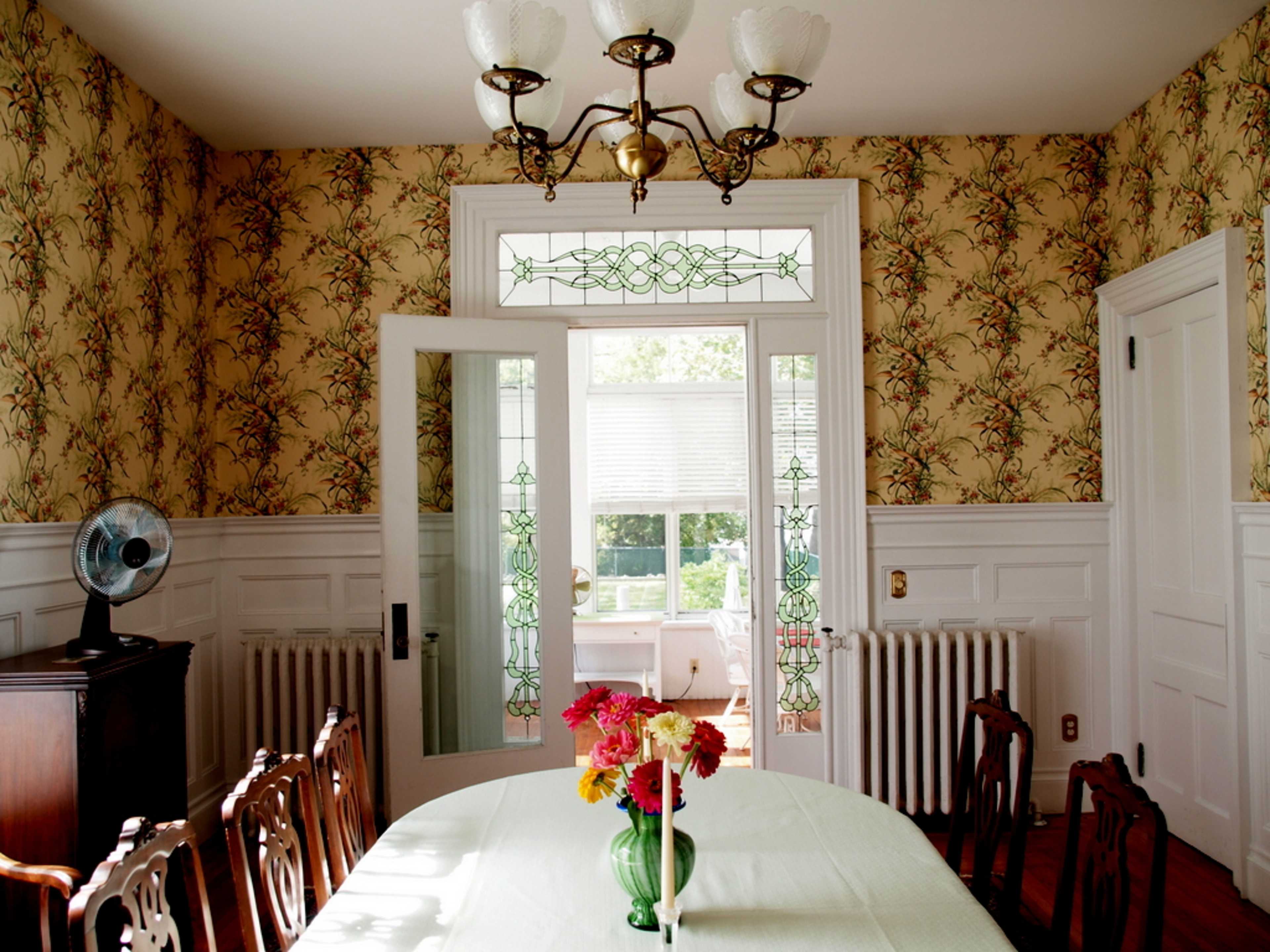 The image shows a dining room with a floral wallpaper, a large table at the center, a vase of flowers, and a door leading to a sunlit area.