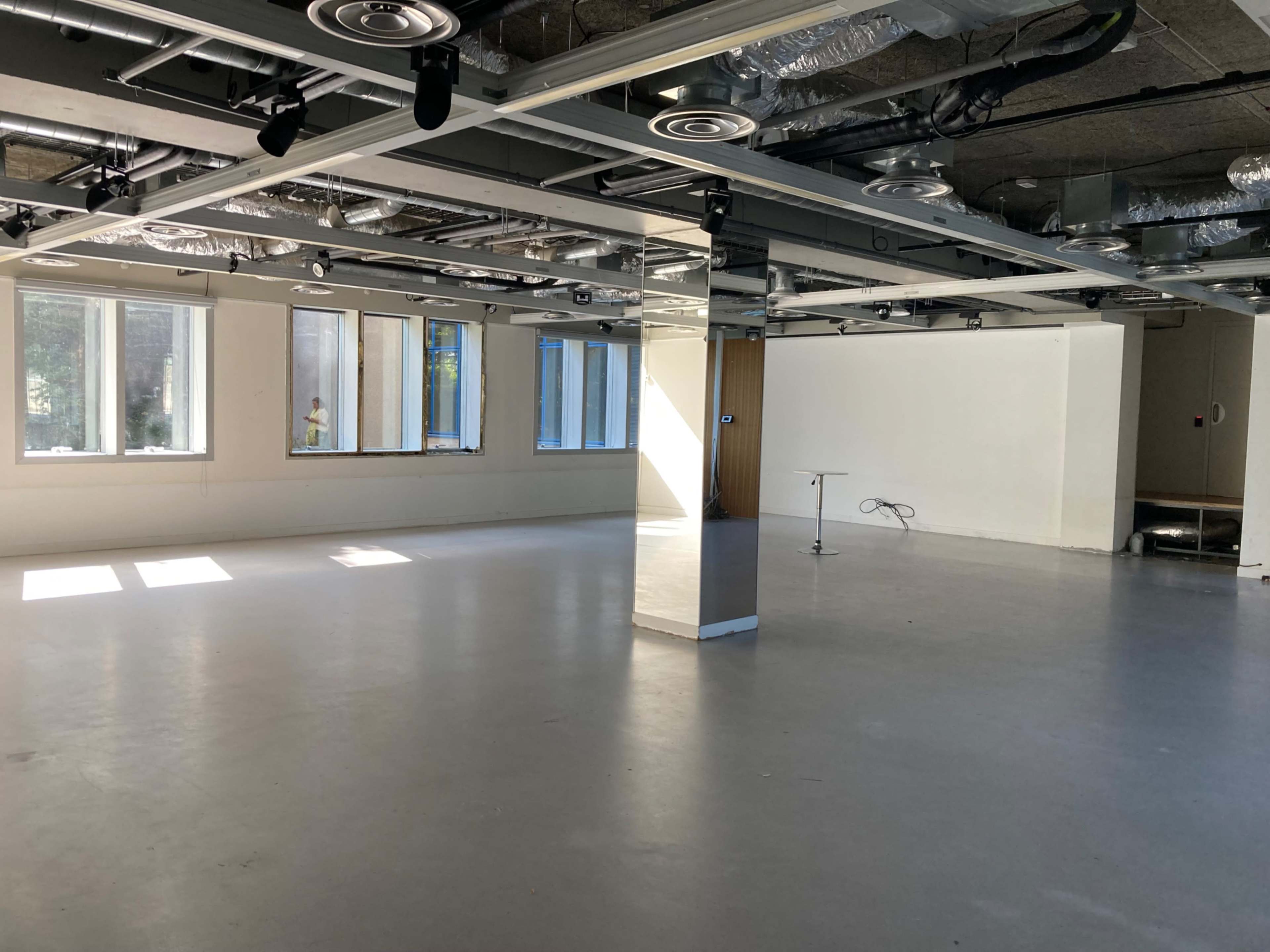The image shows an empty room with large windows, a smooth gray floor, and exposed ductwork on the ceiling.