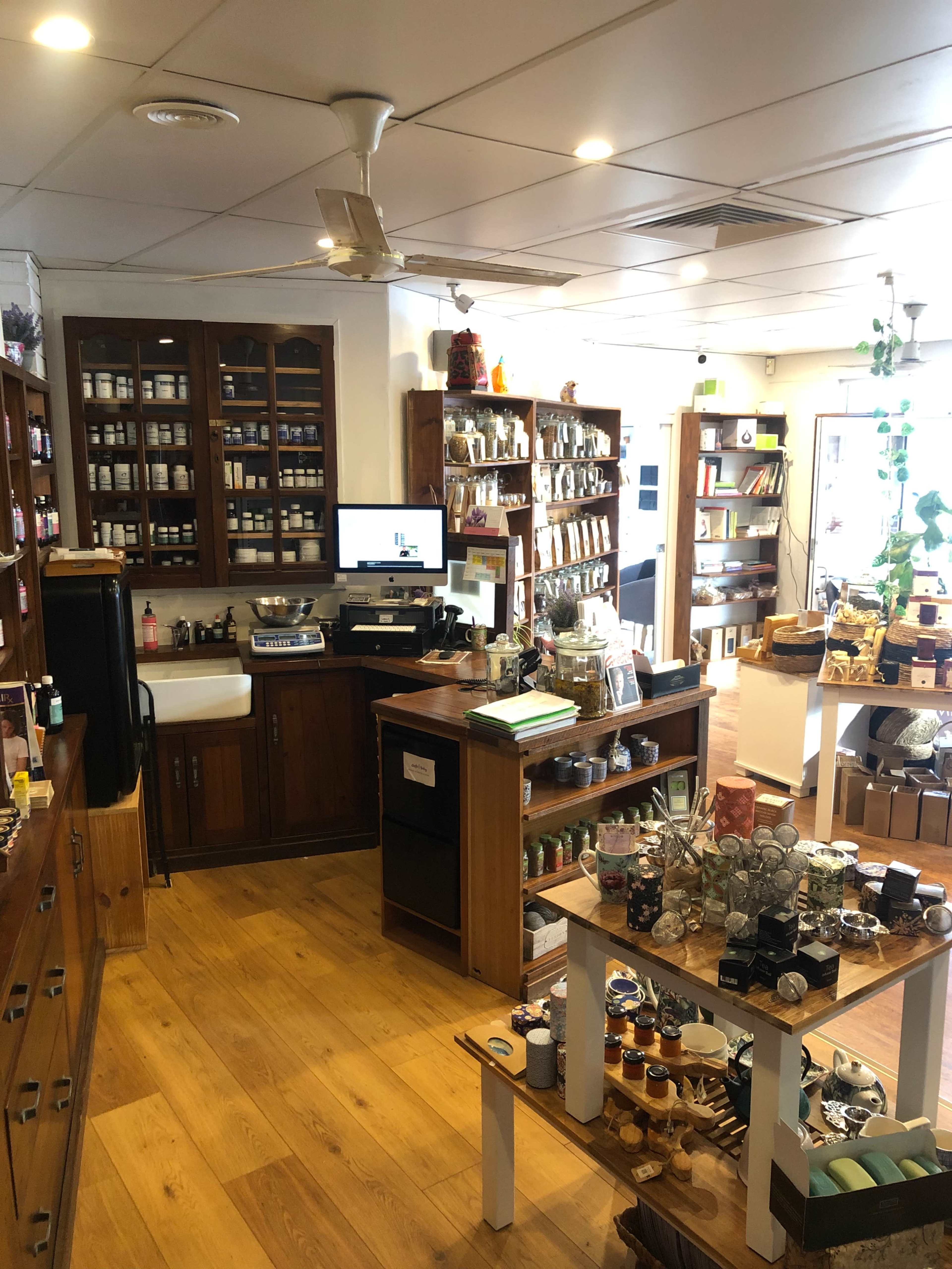 The image shows the interior of a herbal store with wooden shelves filled with jars and boxes, a counter with a computer, and various products displayed on tables.