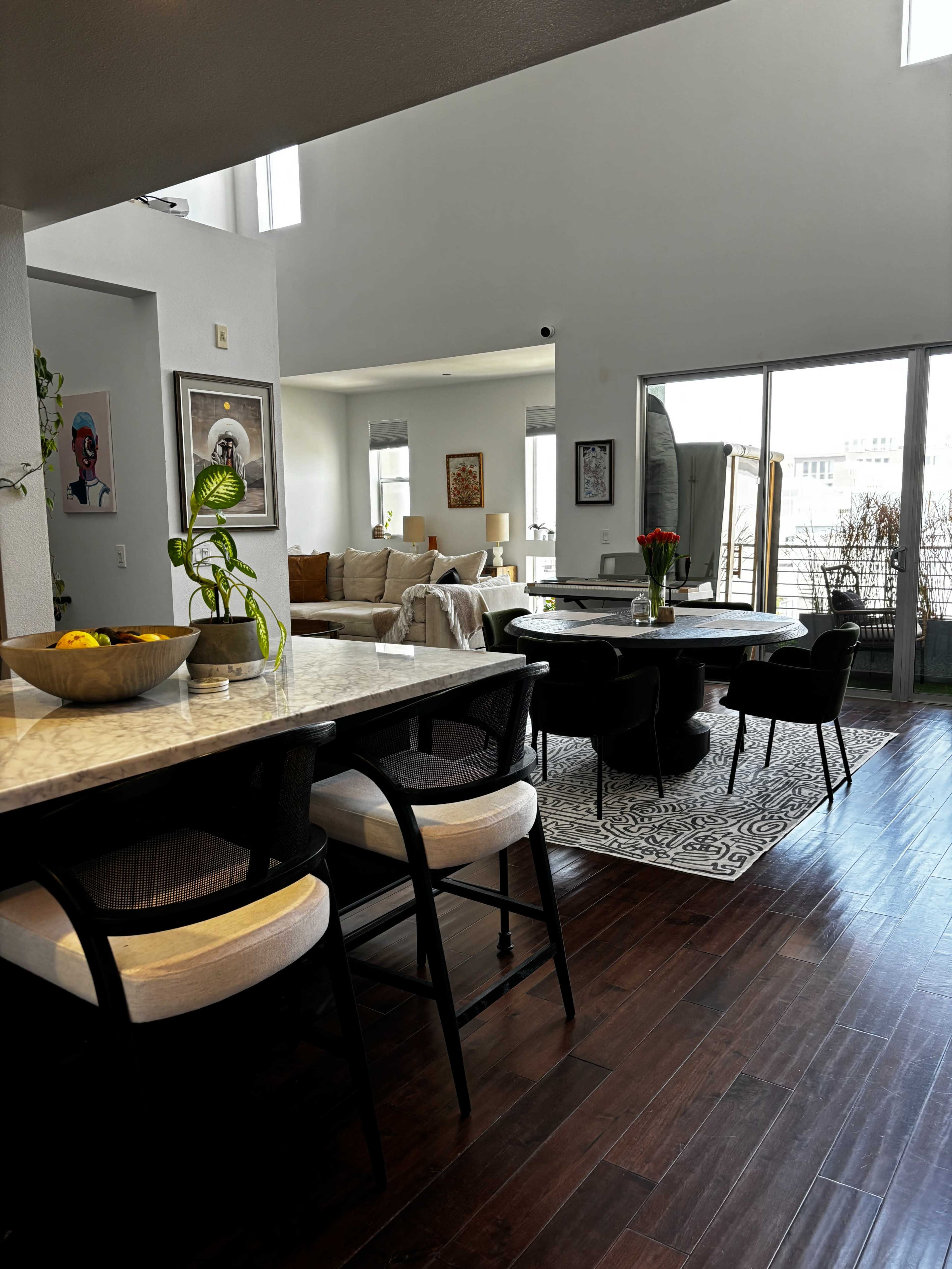 A spacious, modern living area featuring a dining table, bar stools, and large windows allowing natural light.