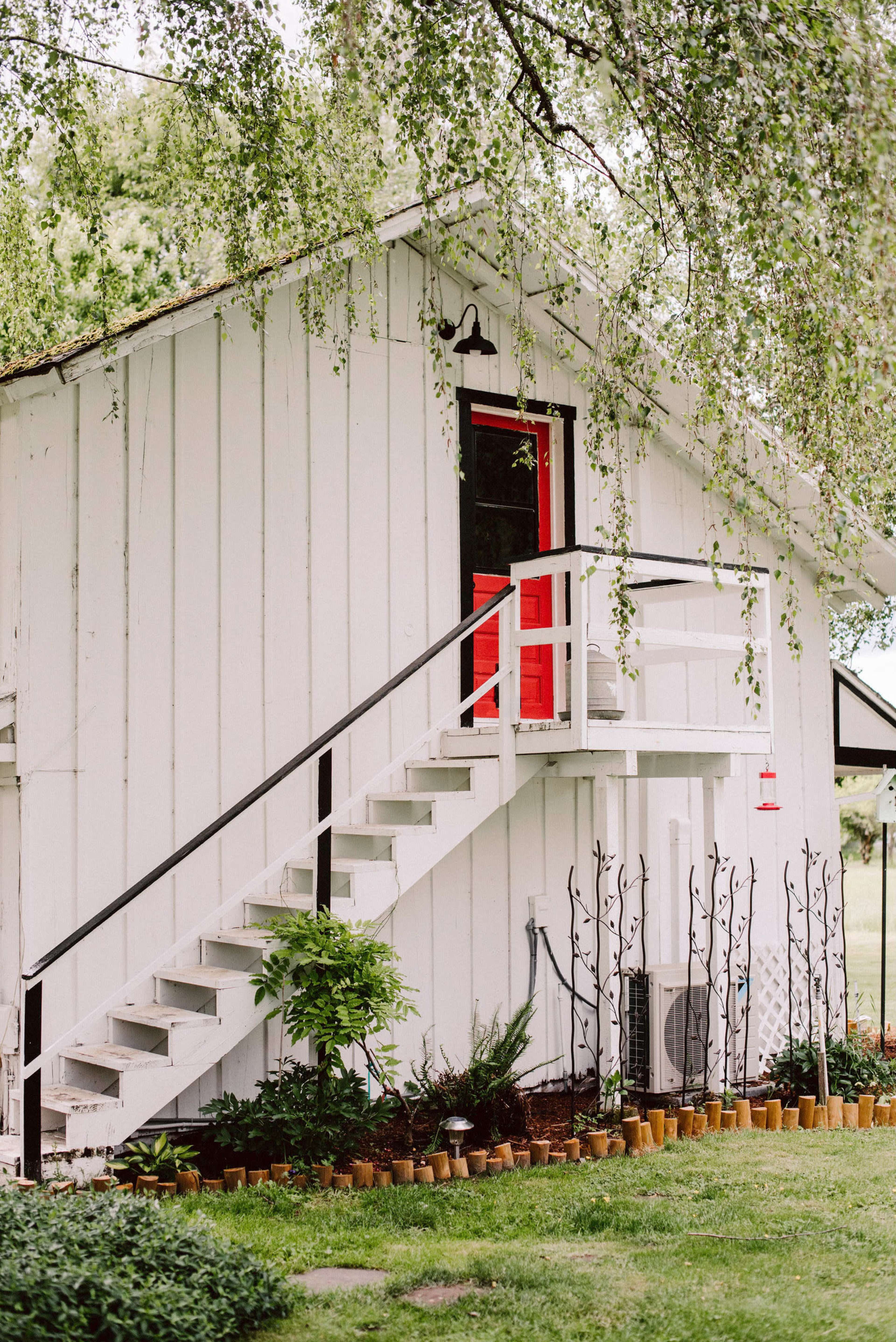 A white wooden house features a red door and a staircase leading to it, surrounded by a garden with potted plants.