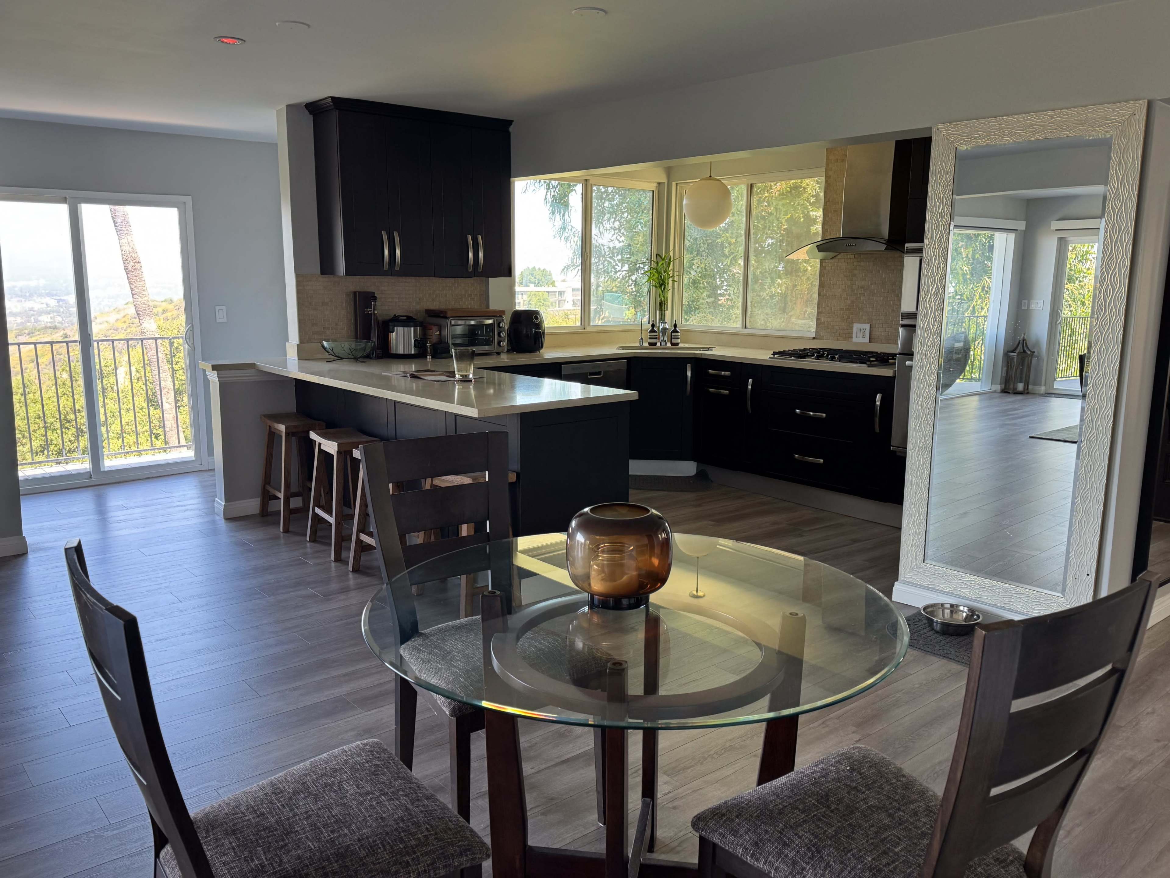 A modern kitchen and dining area with a round glass table, surrounded by wooden chairs, and large windows offering a view of the outdoors.